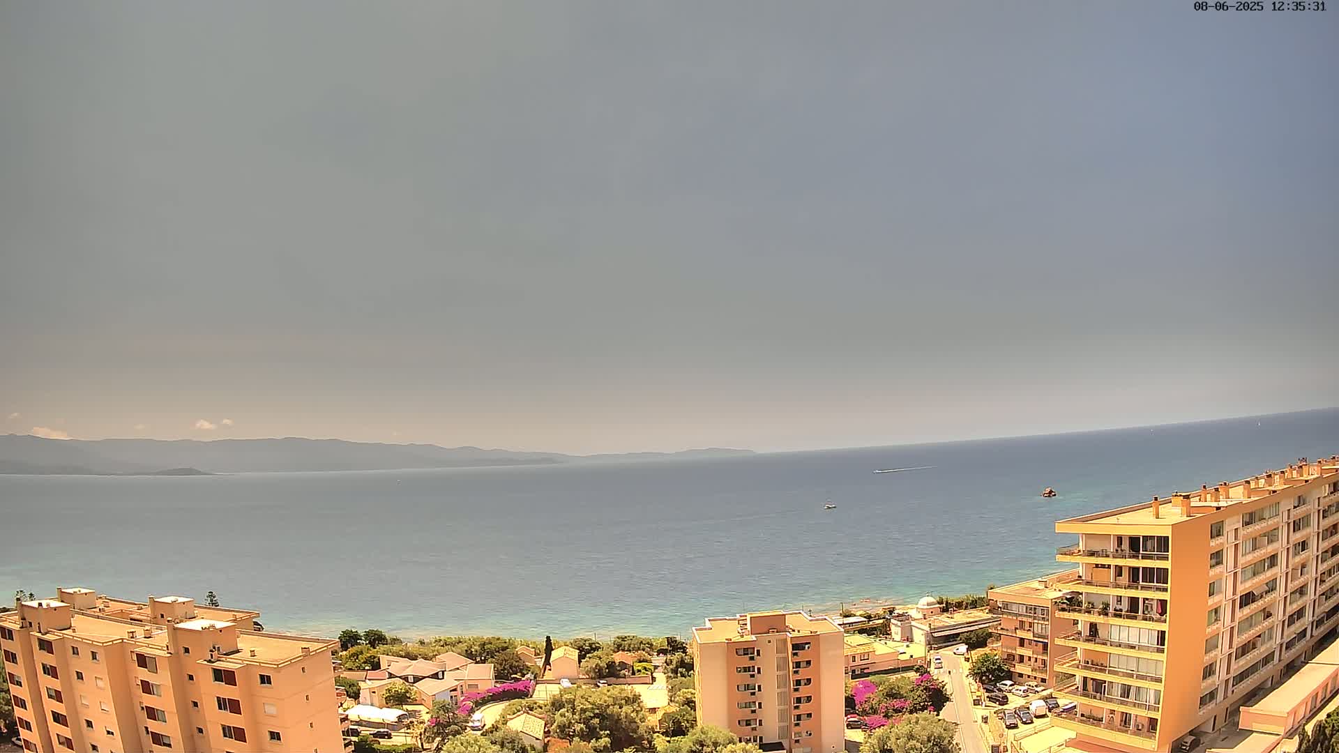 An expansive view of a calm sea under a hazy sky overlooks a coastal town with several multi-story buildings.