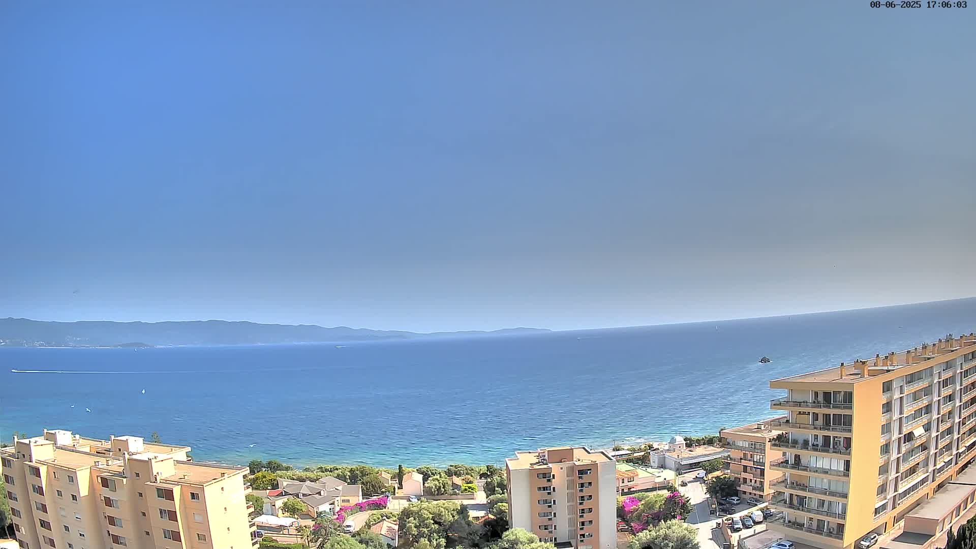 A clear, sunny day overlooks a calm blue sea, with several buildings and a small coastal town visible in the foreground and mountains in the distance.