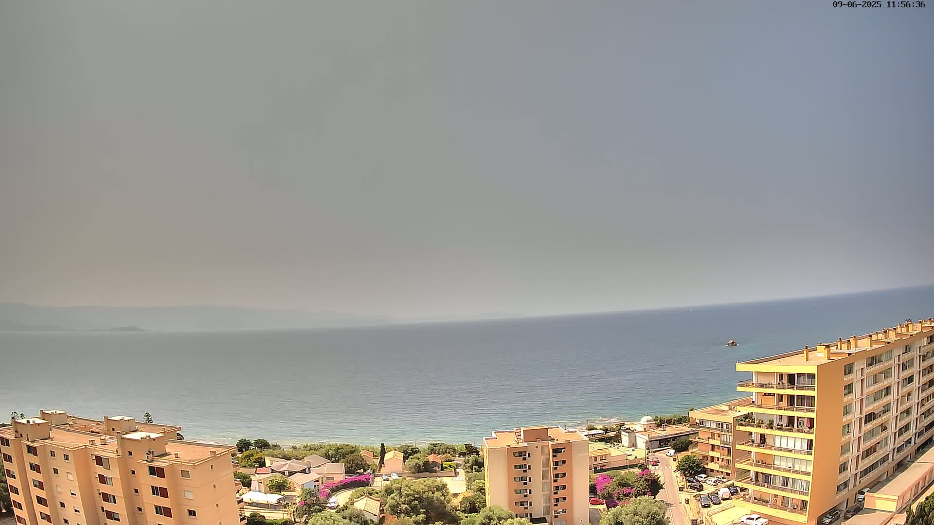 An aerial view shows a hazy coastal scene with calm water, a shoreline with buildings and vegetation, and several multi-story apartment buildings.