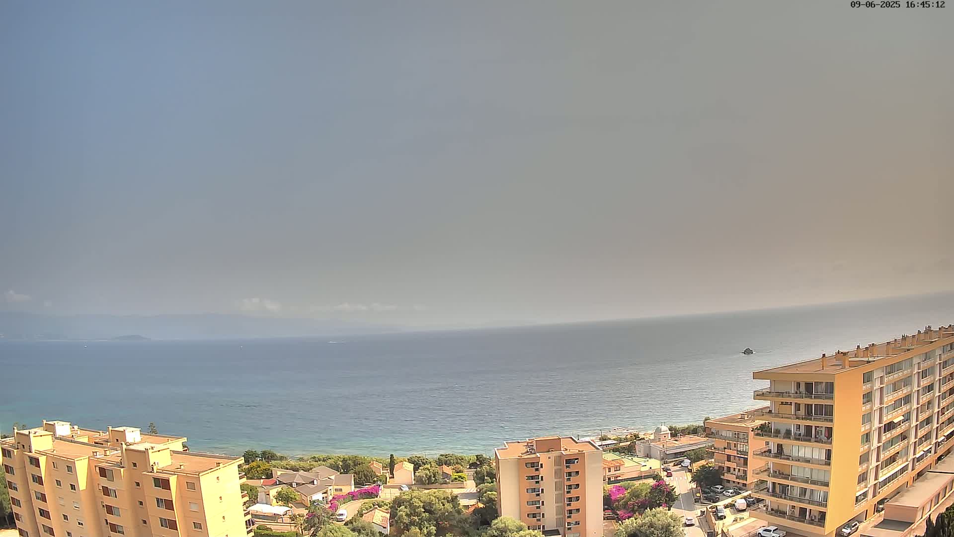 A hazy, sunny day overlooks a calm sea from an elevated vantage point showing a residential area with several apartment buildings near the coast.