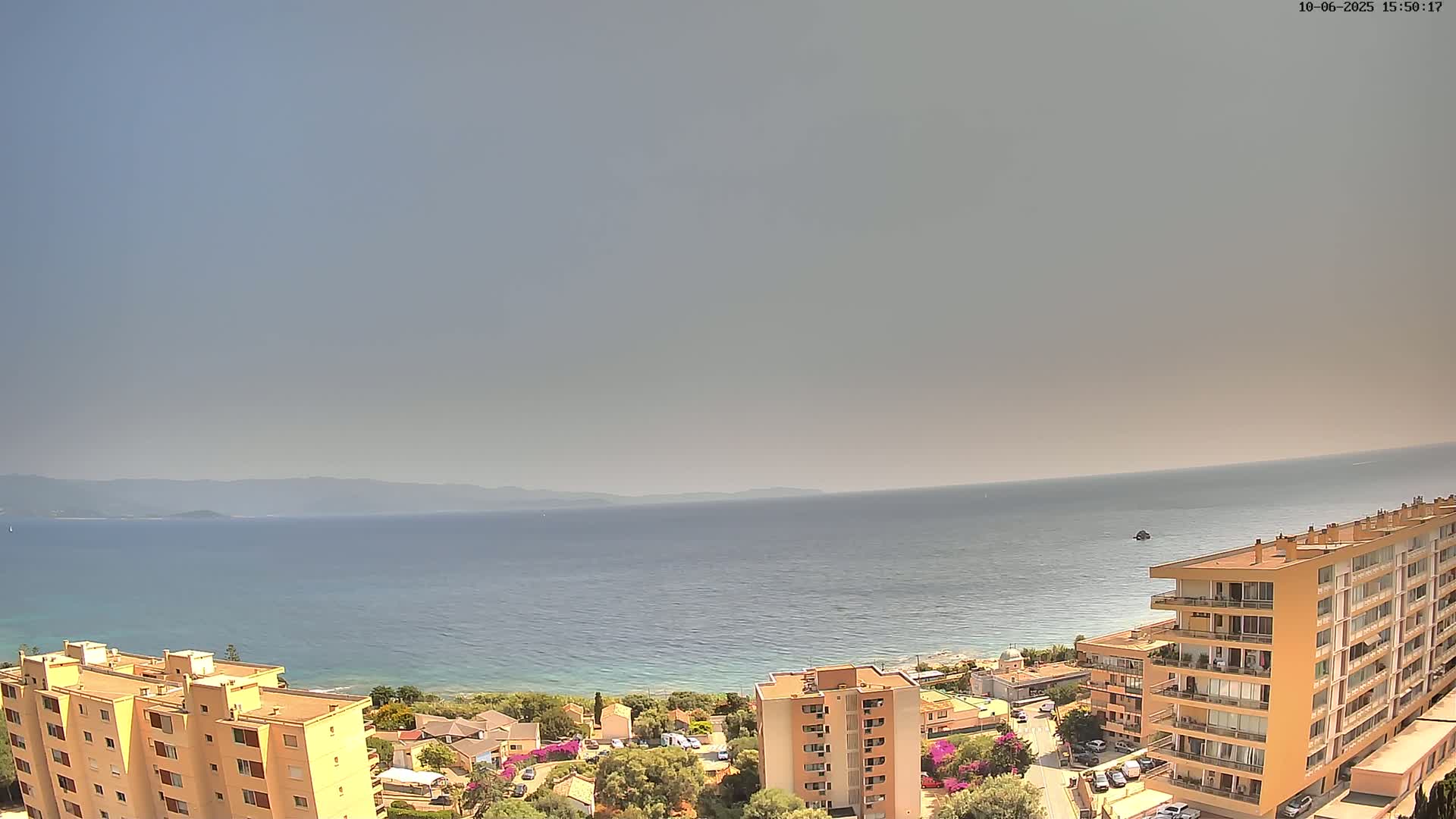 A hazy, sunny day overlooks a calm sea, with multiple apartment buildings and a coastline visible in the foreground.