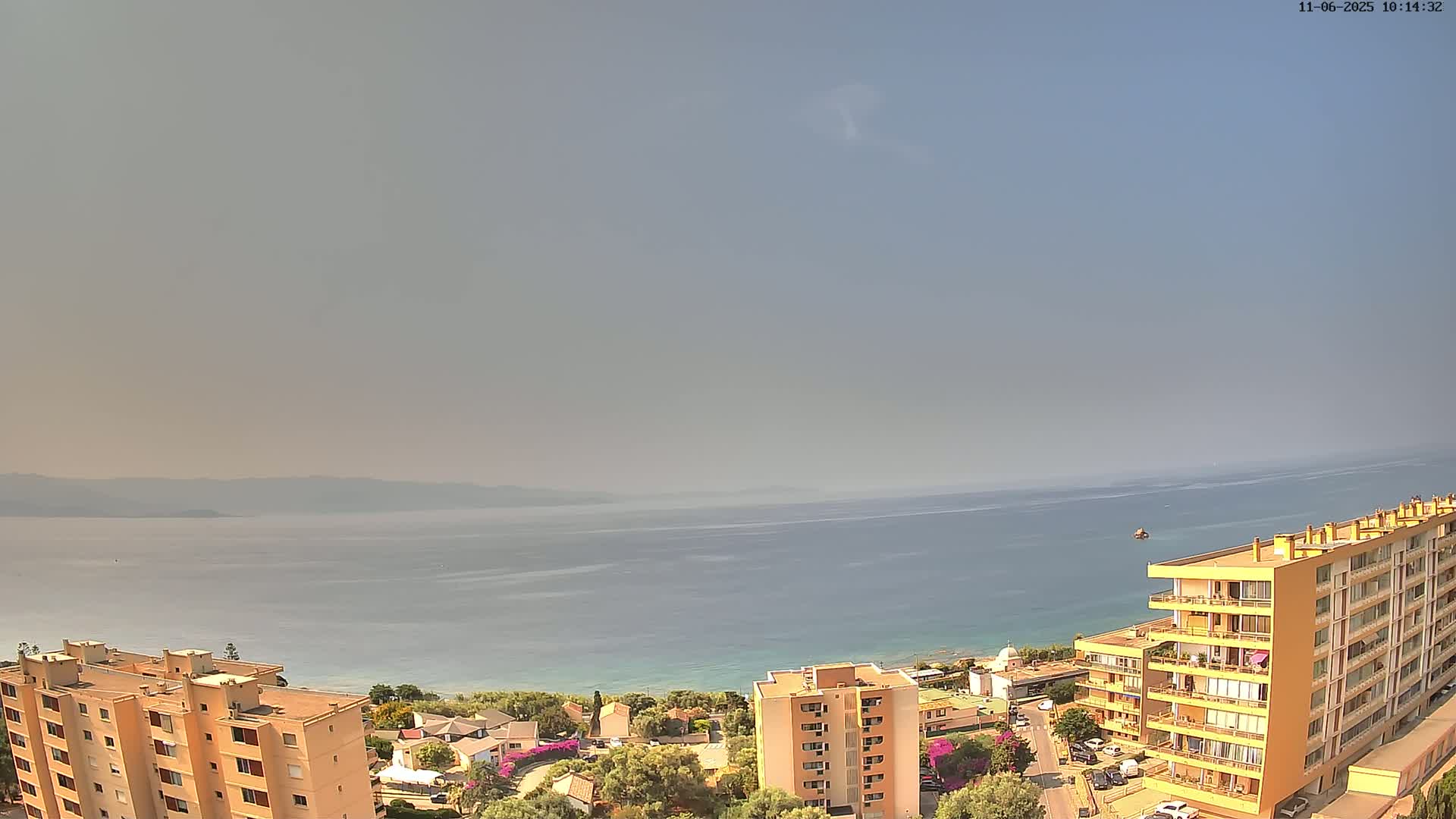 A hazy, sunny day overlooks a calm ocean, with multiple apartment buildings and surrounding vegetation visible in the foreground.