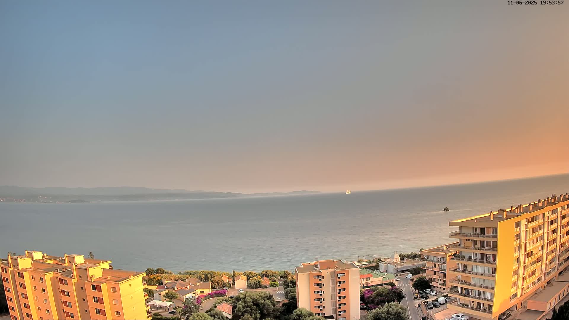 A calm sea at sunset is viewed from above, showing a coastal town with apartment buildings and a distant vessel.