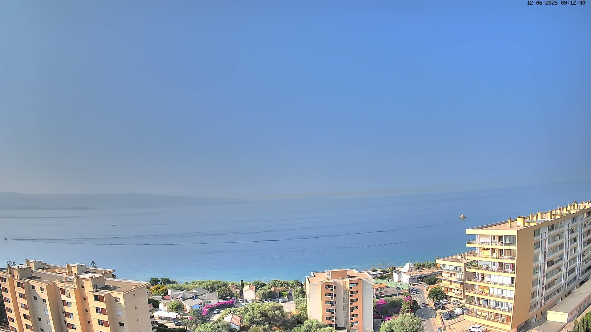 A clear, sunny day overlooks a calm blue sea and coastline, with several apartment buildings visible in the foreground.