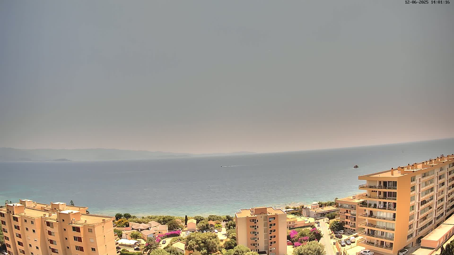A hazy, sunny day overlooks a calm sea, with distant mountains and several multi-story buildings on a hillside near the coast.
