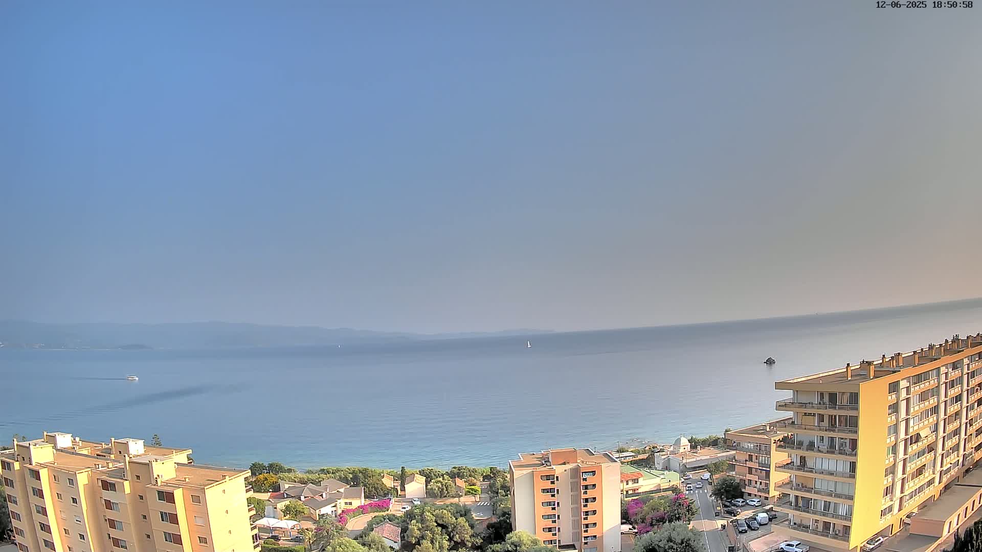 A calm sea stretches to a hazy horizon under a clear sky, overlooking a coastal town with multi-story buildings.