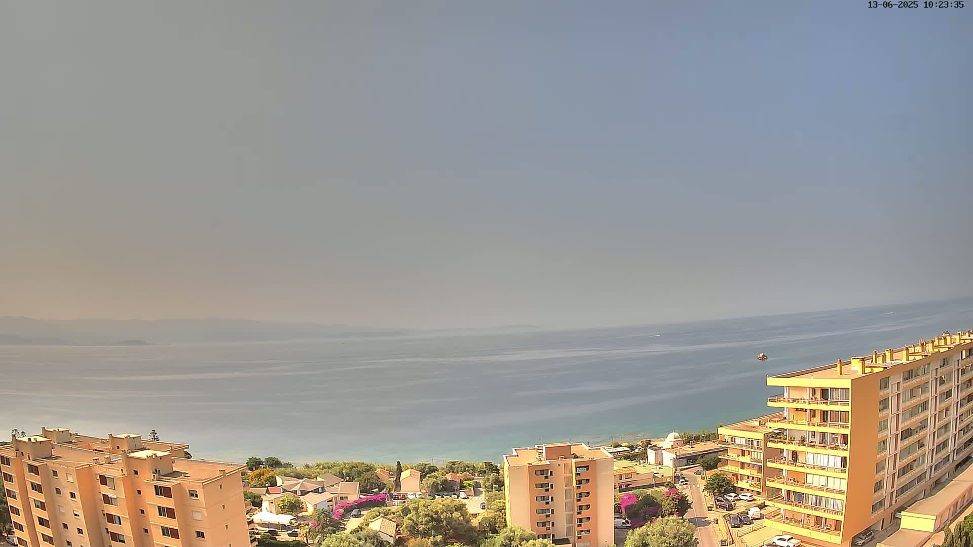 An aerial view shows hazy weather over a calm sea, with multiple apartment buildings and other structures situated on a hillside overlooking the water.
