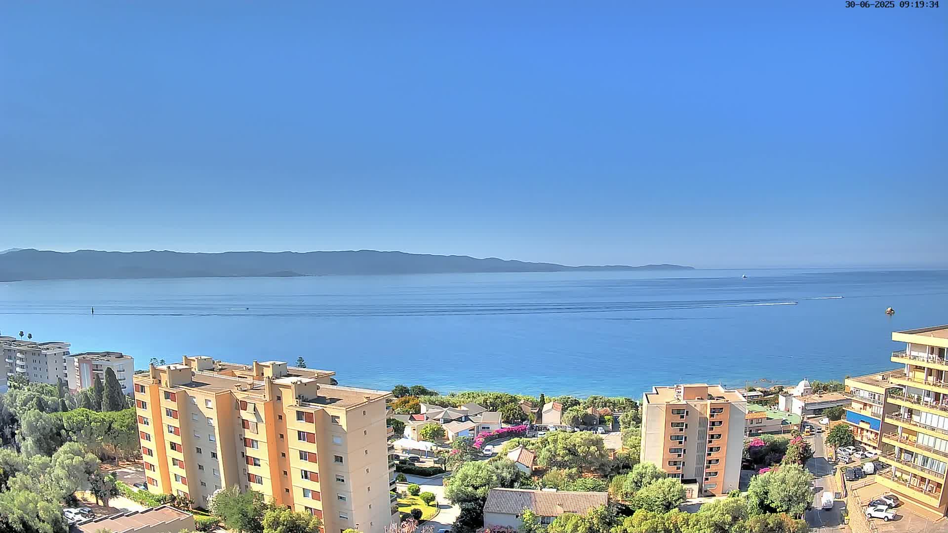 A clear, sunny day overlooks a calm blue sea with distant mountains, a shoreline of buildings and vegetation, and several boats visible on the water.