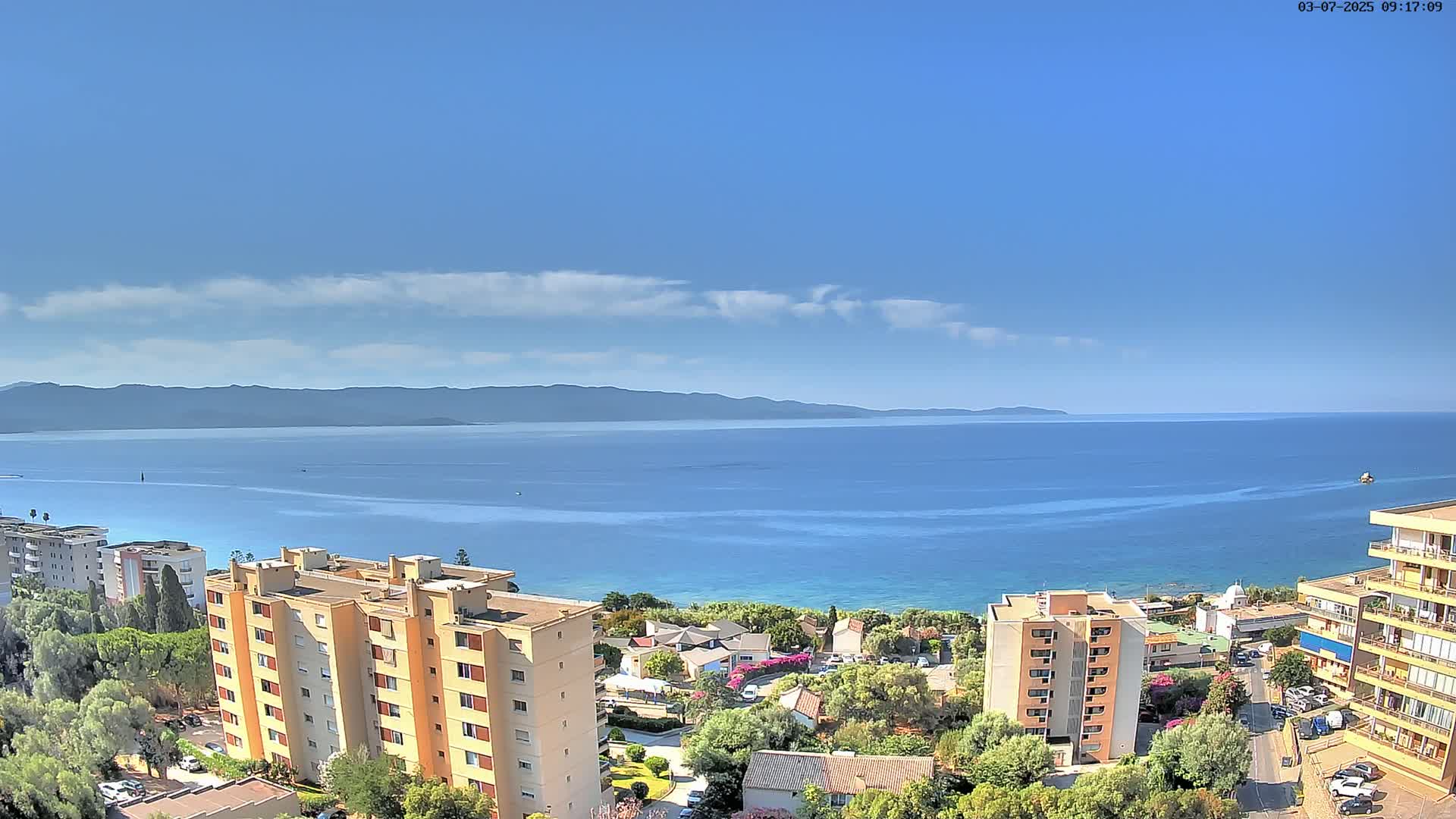 A coastal scene on a partly sunny day shows a calm blue sea, mountains in the distance, and several apartment buildings nestled amongst green trees.