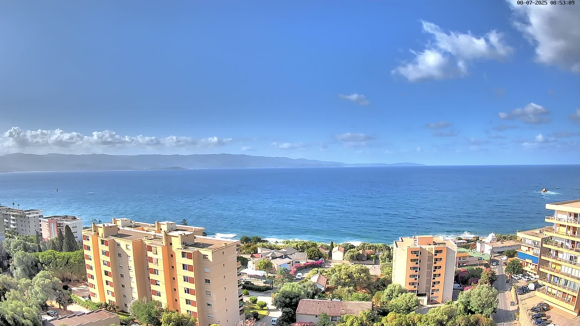 A partly cloudy day overlooks a calm ocean with distant mountains, featuring several multi-story buildings nestled along a coastline with green vegetation.