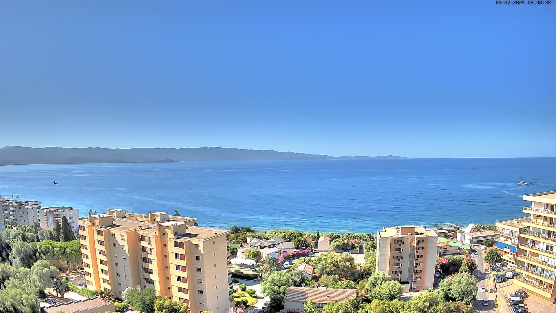 A clear, sunny day shows a coastal view of several apartment buildings and green vegetation overlooking a calm blue sea and distant mountains.
