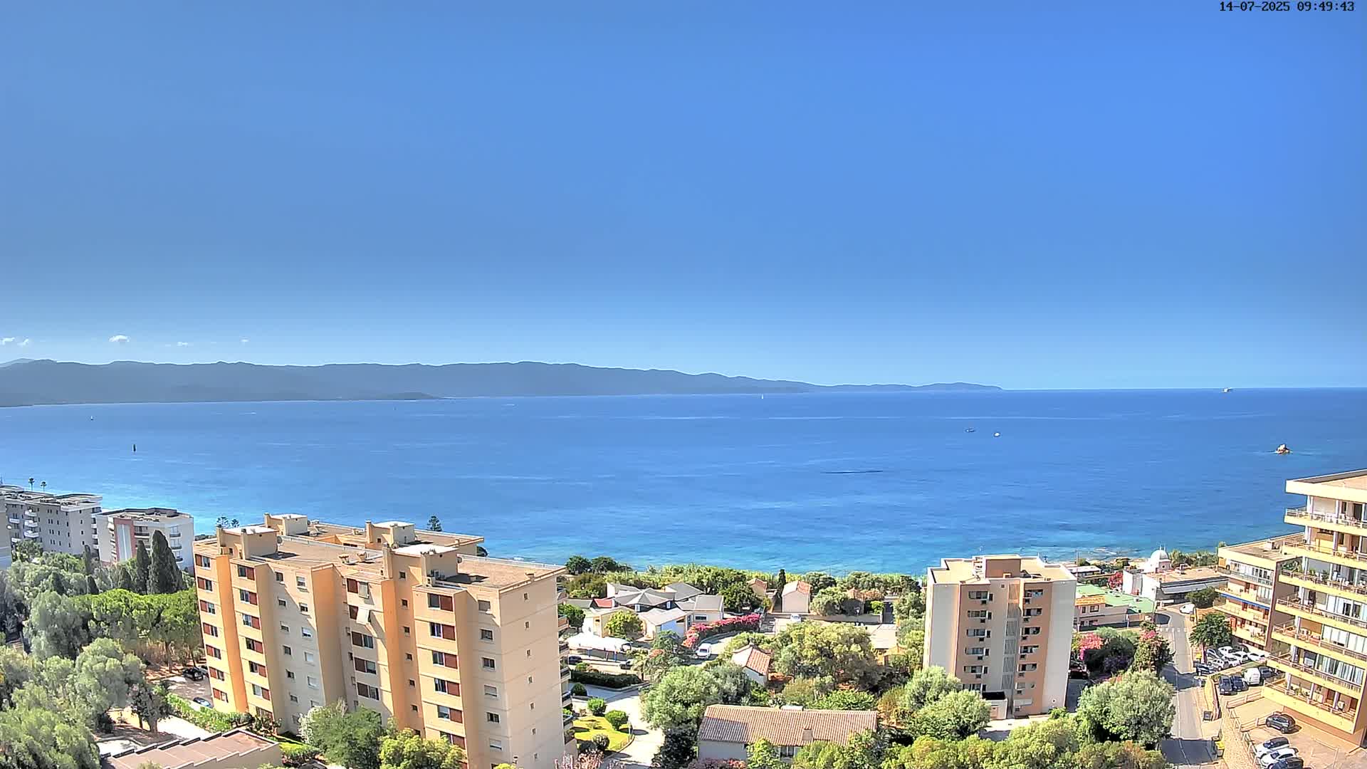 A sunny day overlooking a calm blue sea, with several multi-story buildings and green vegetation situated along the coastline, and mountains visible in the distance.