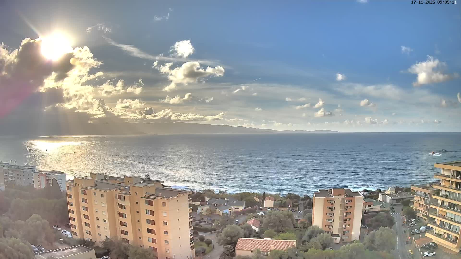 On a bright and partly cloudy day, the sun intensely reflects on the ocean surface, illuminating a coastal town featuring residential buildings and a winding shore, with distant mountains under a sky dotted with white clouds.