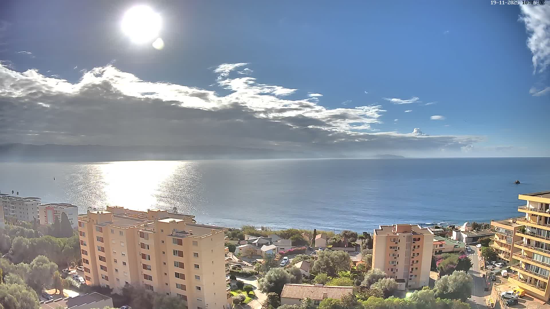 On a bright, sunny day with some clouds, a high-angle view reveals multi-story buildings and green trees along a coastline, overlooking a sparkling blue sea with distant land visible on the horizon.