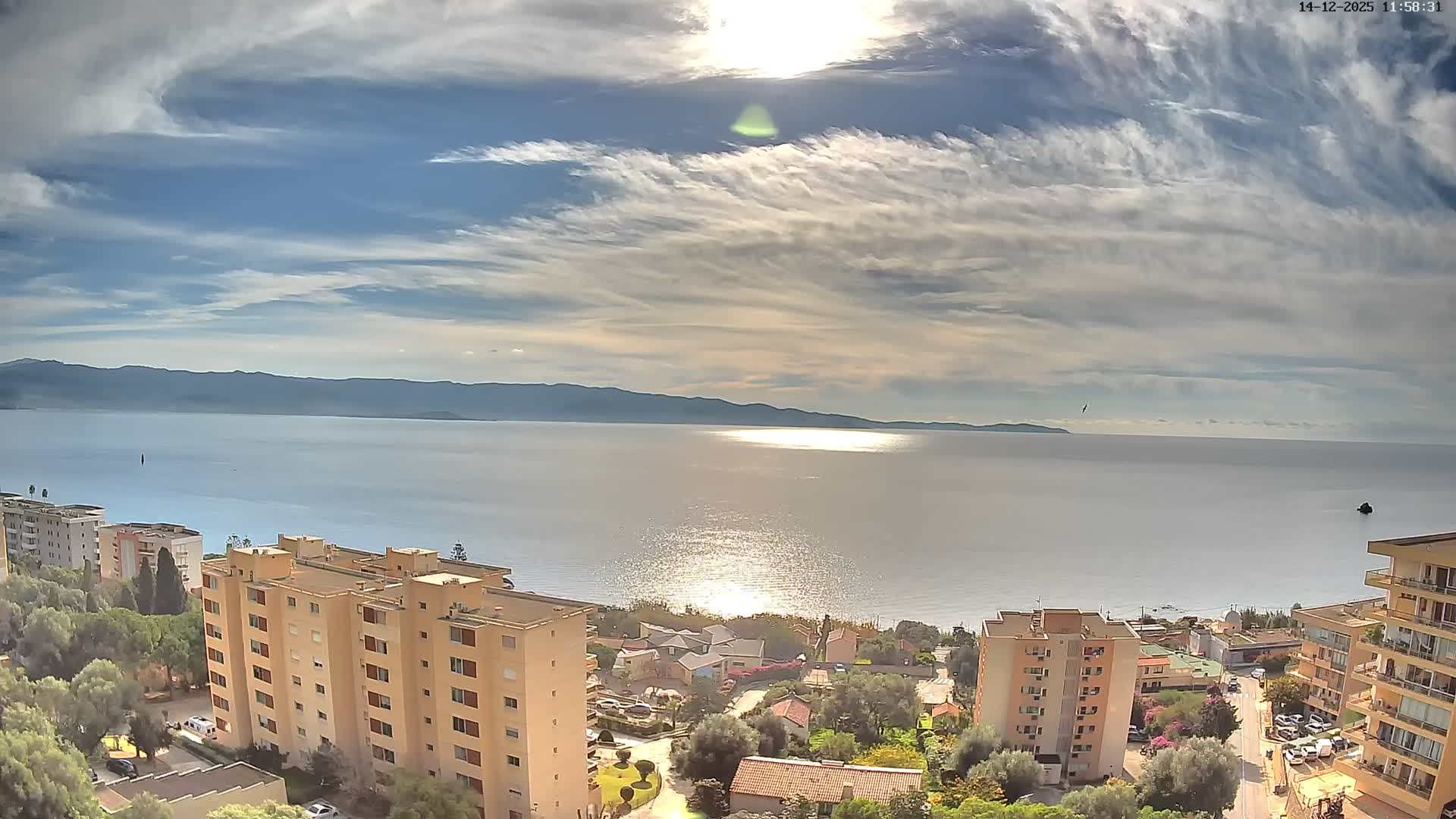 On a bright, sunny day with some clouds, a high-angle view reveals multi-story buildings and green trees along a coastline, overlooking a sparkling blue sea with distant land visible on the horizon.