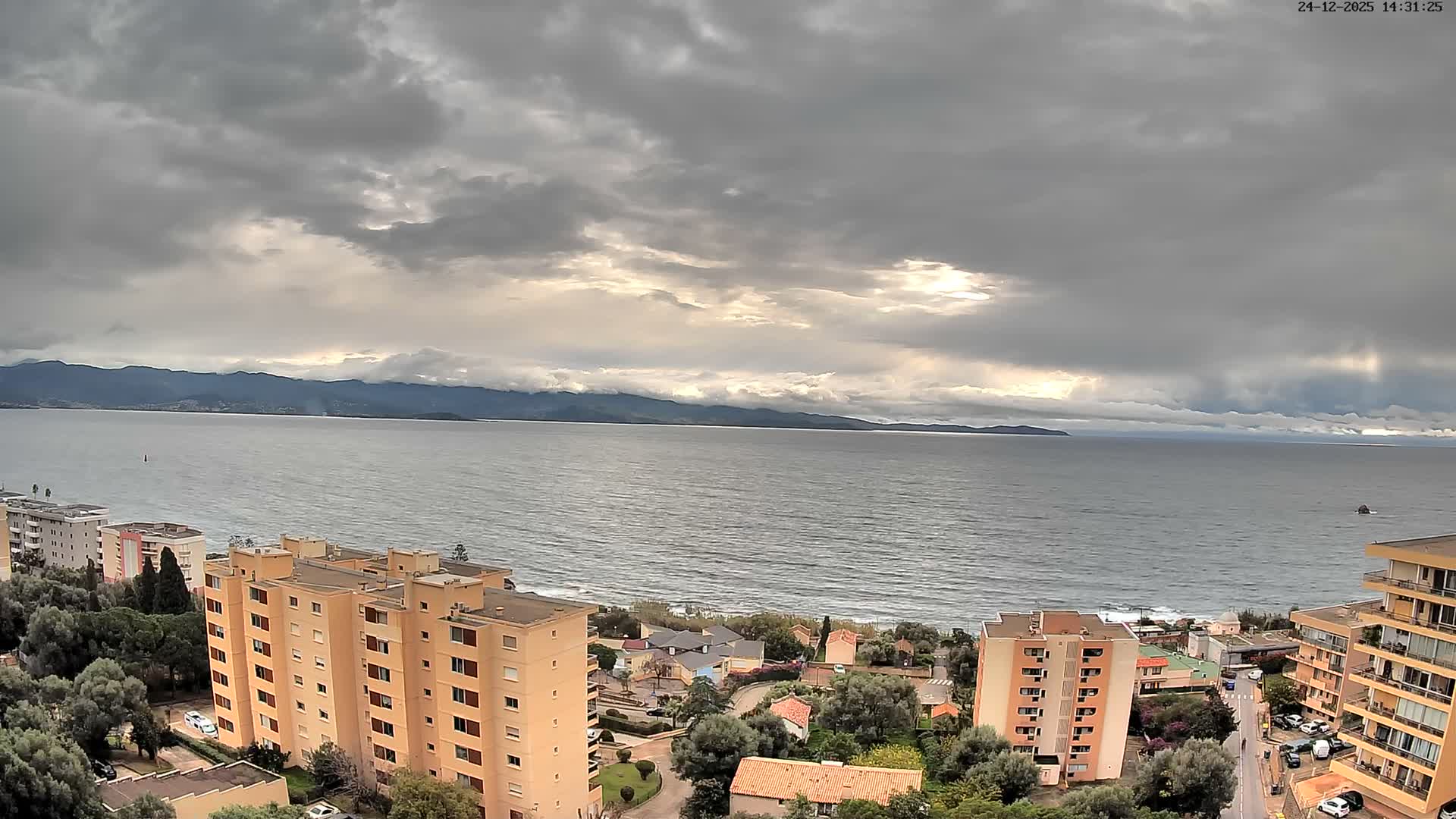 On a bright, sunny day with some clouds, a high-angle view reveals multi-story buildings and green trees along a coastline, overlooking a sparkling blue sea with distant land visible on the horizon.