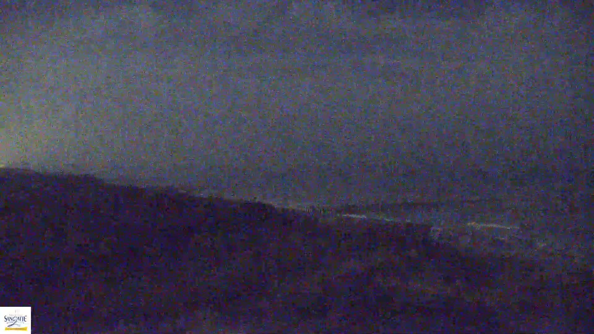 A dark, overcast sky hangs over a hilly landscape at night, with a body of water visible in the distance.