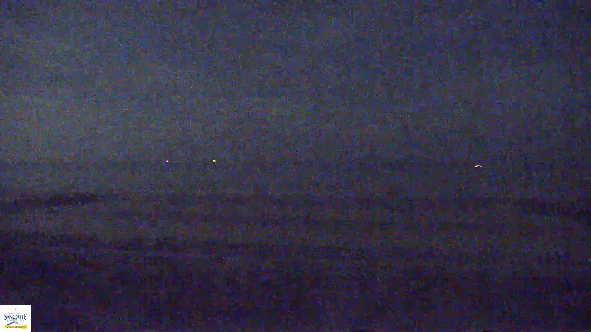 A dark, nighttime scene shows a flat, dark landscape with a few distant, faint lights on the horizon under a cloudy, dark sky.