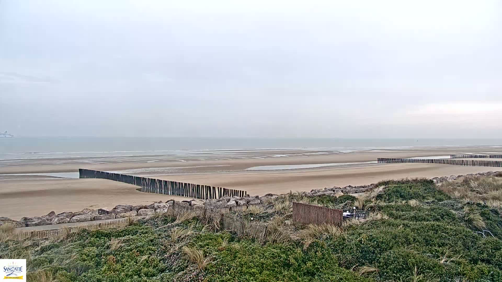 A wide sandy beach with low wooden barriers stretches to a calm, gray sea under an overcast sky.