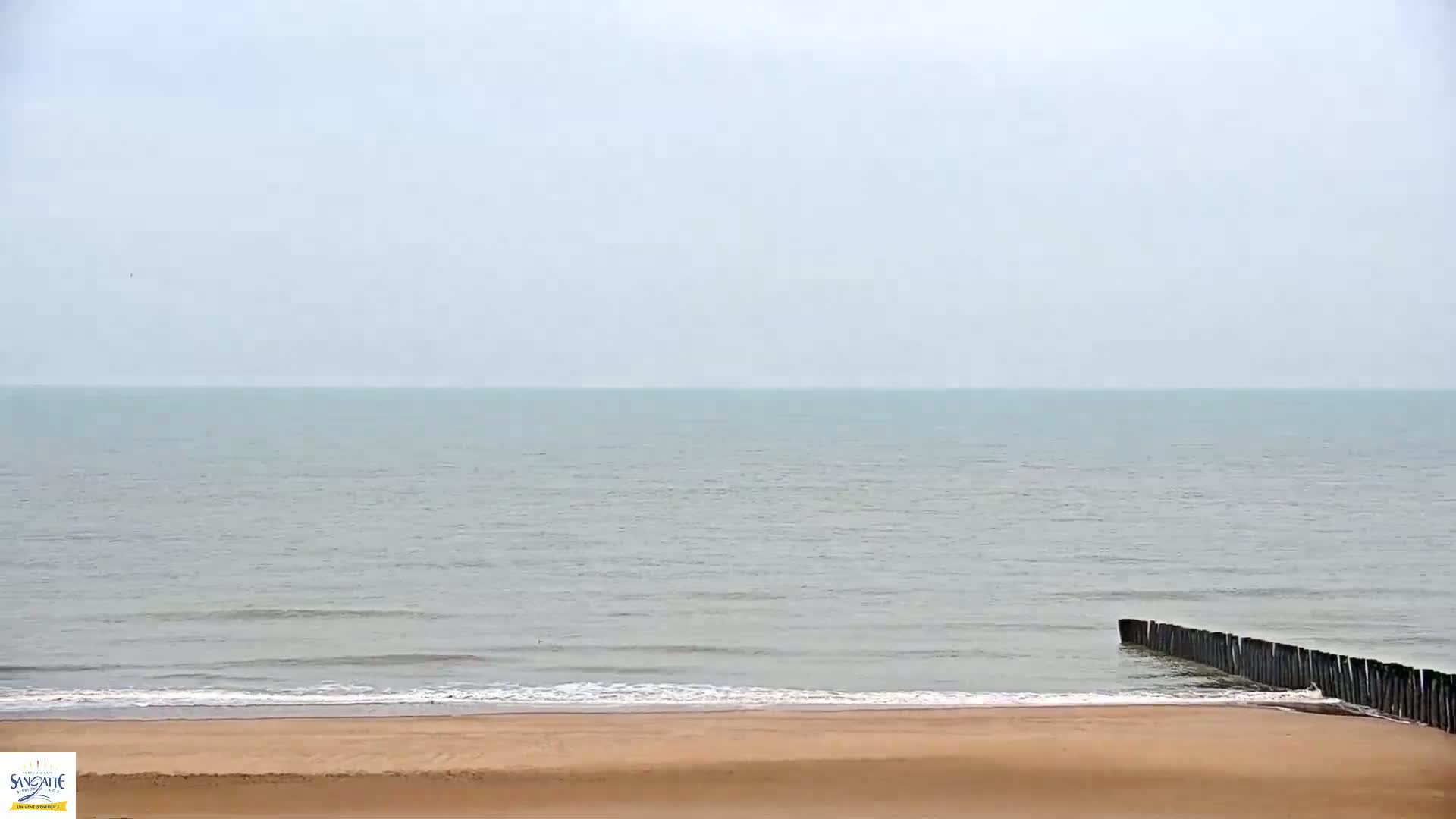 A calm, light-grey sea meets a sandy beach where a short, dark wooden breakwater juts into the water.