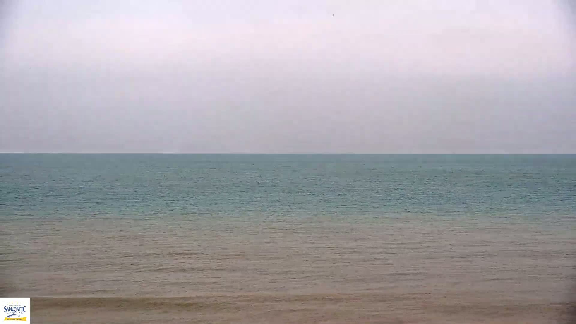 A calm, overcast day at the beach shows a mostly flat ocean with a subtle color gradient from teal to tan near the shore.