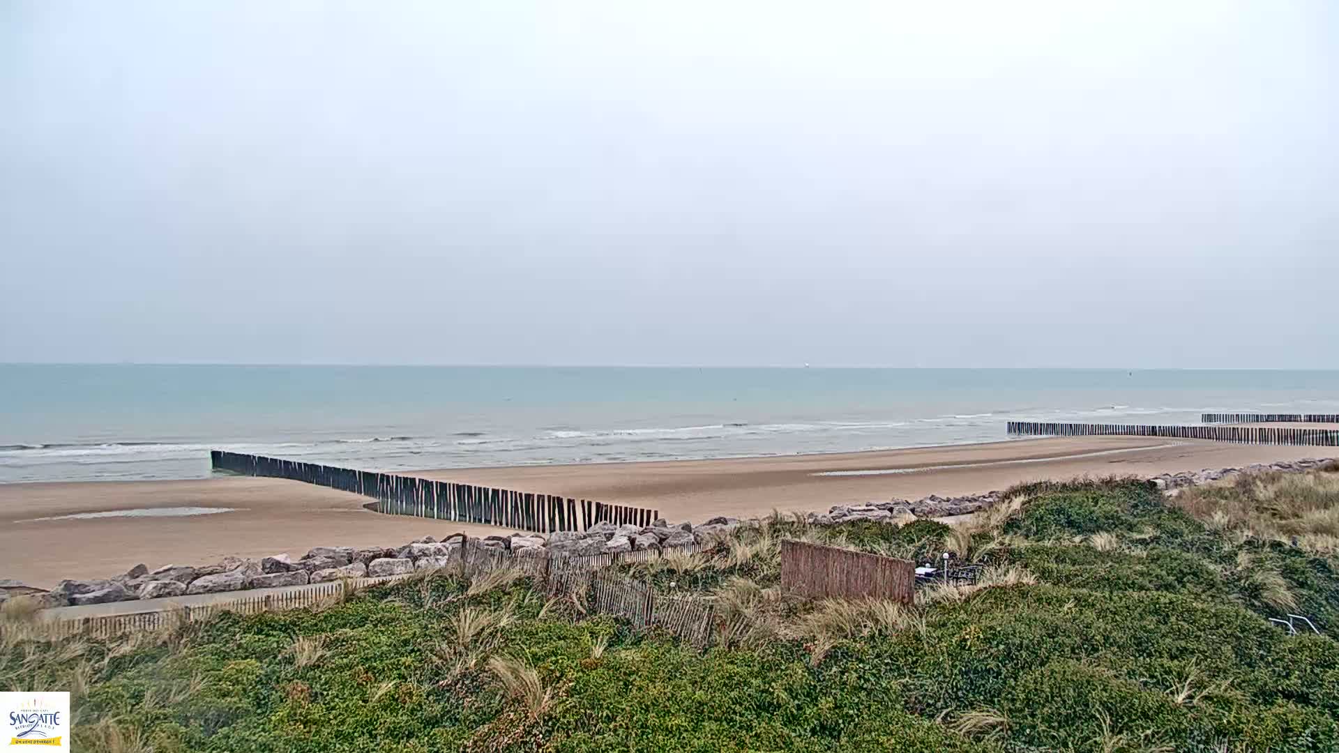 A calm, light-blue ocean meets a sandy beach protected by wooden barriers, all under a cloudy sky.