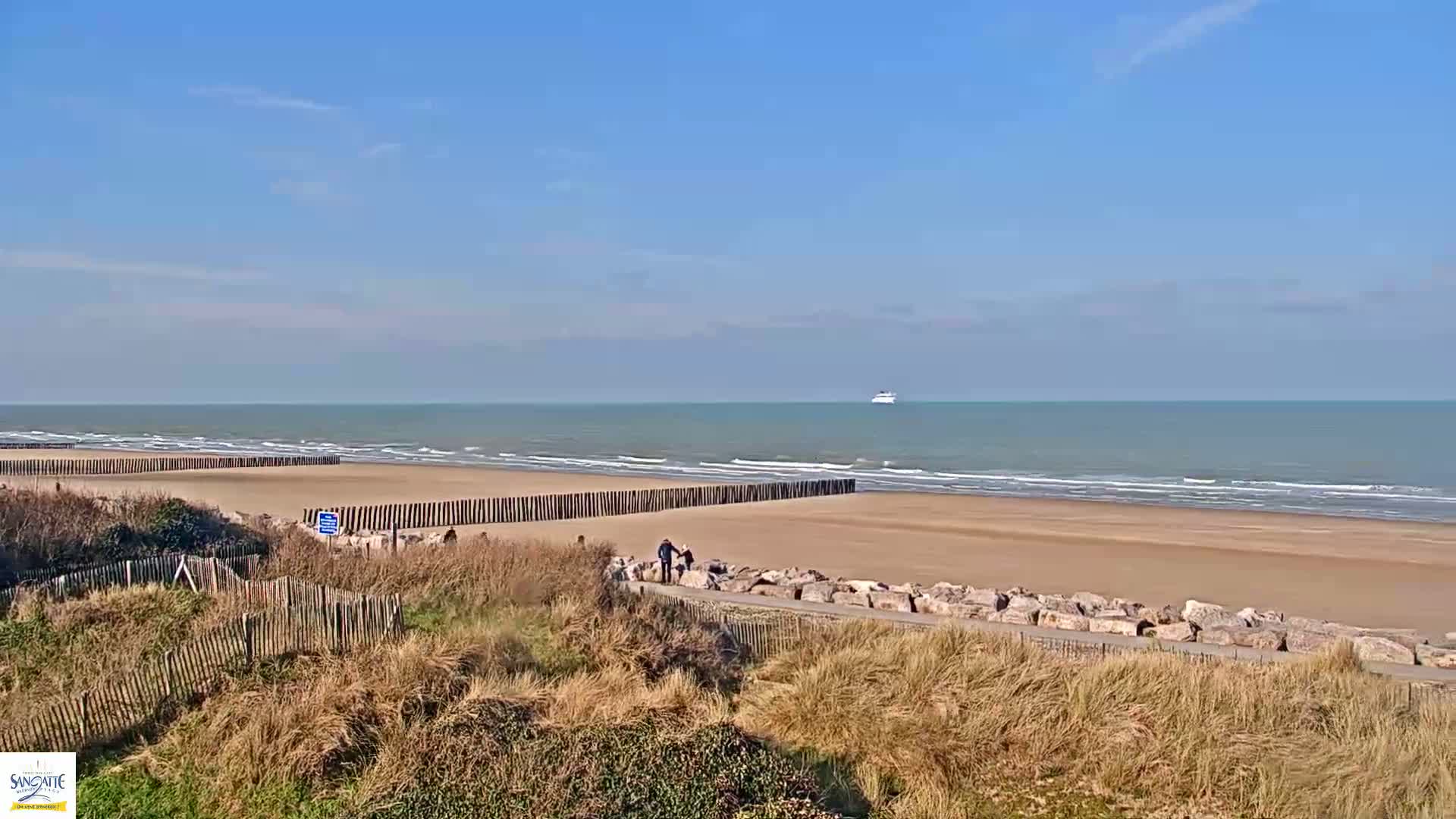 A sandy beach with a calm ocean under a clear blue sky, featuring a few people walking near wooden posts and rock barriers.