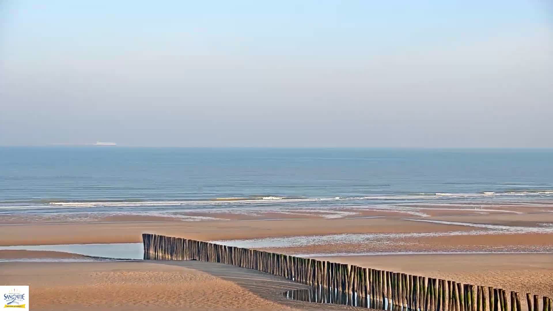 A sandy beach with a low wooden fence stretches along a calm ocean under a hazy sky.