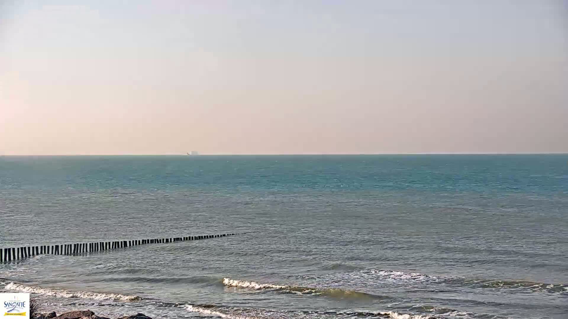 A calm sea with a hazy sky stretches to the horizon where a distant ship is visible, and small waves lap against a shoreline protected by a row of wooden posts.