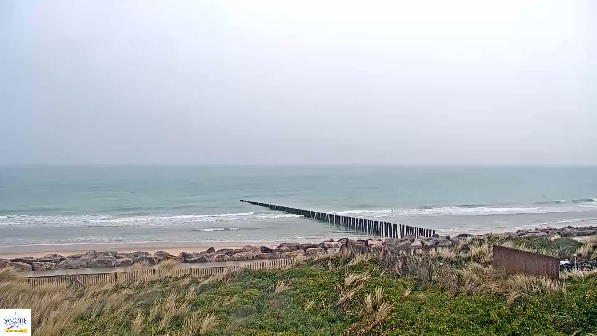 A long, dark structure extends from the shore into a light teal ocean with small waves on an overcast day.