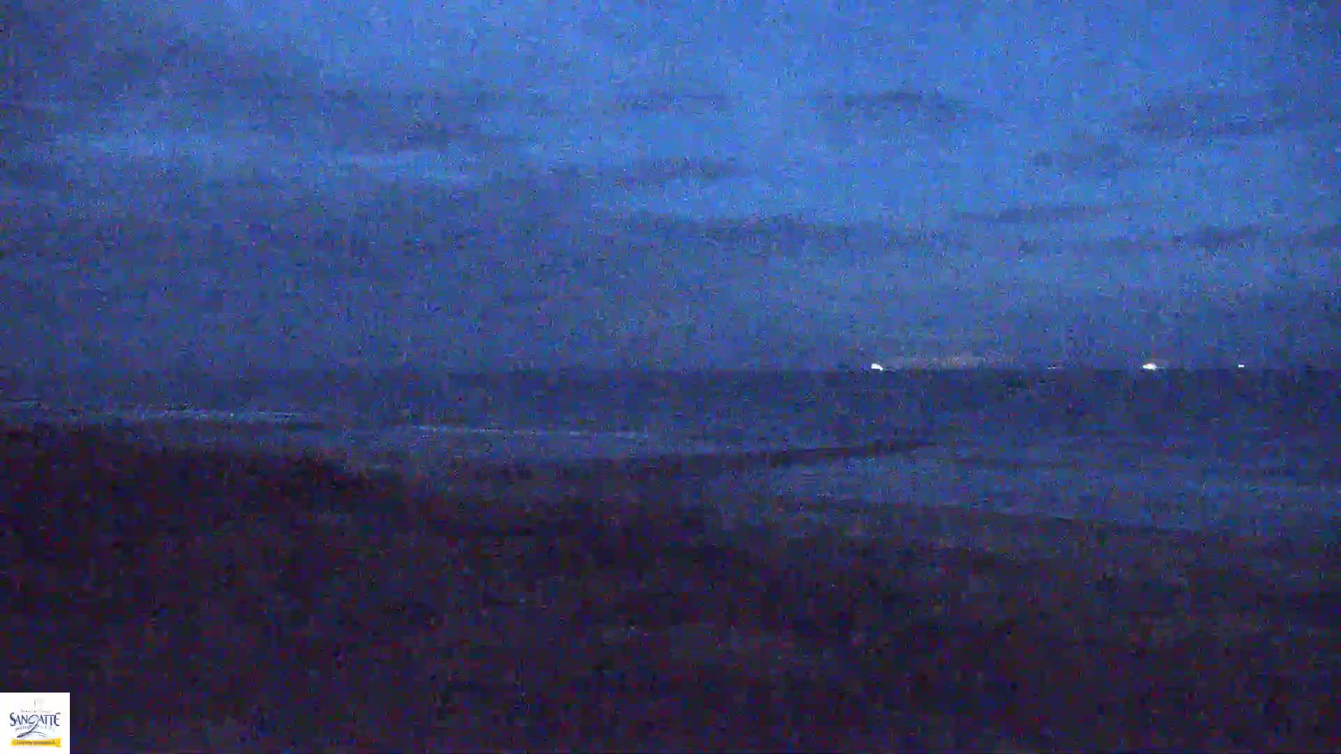A dark, mostly cloudy night scene shows a dark ocean with faint lights on the horizon and a dark shoreline in the foreground.