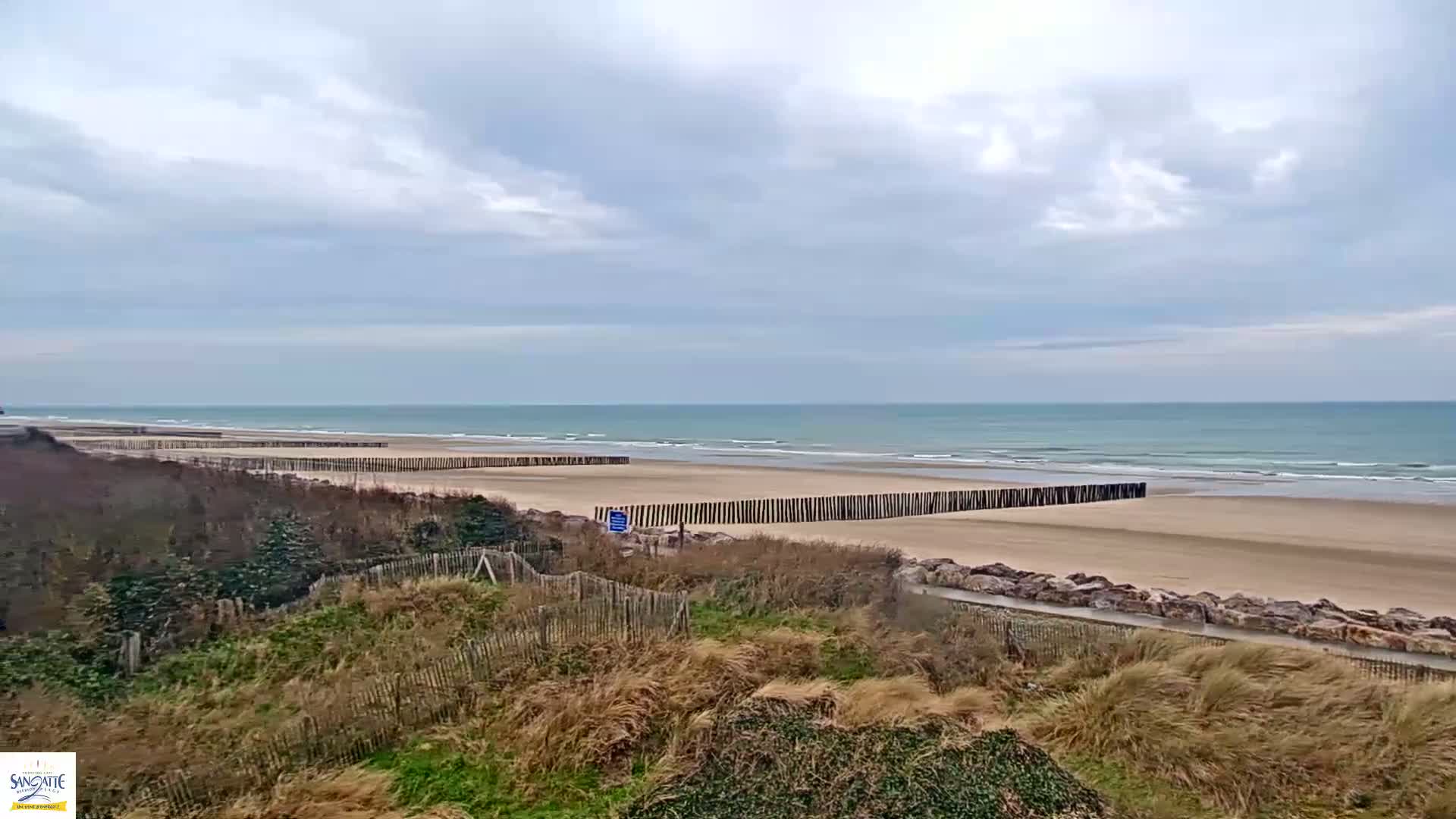 A sandy beach with a calm ocean under a cloudy sky is protected by wooden barriers and fenced dunes.