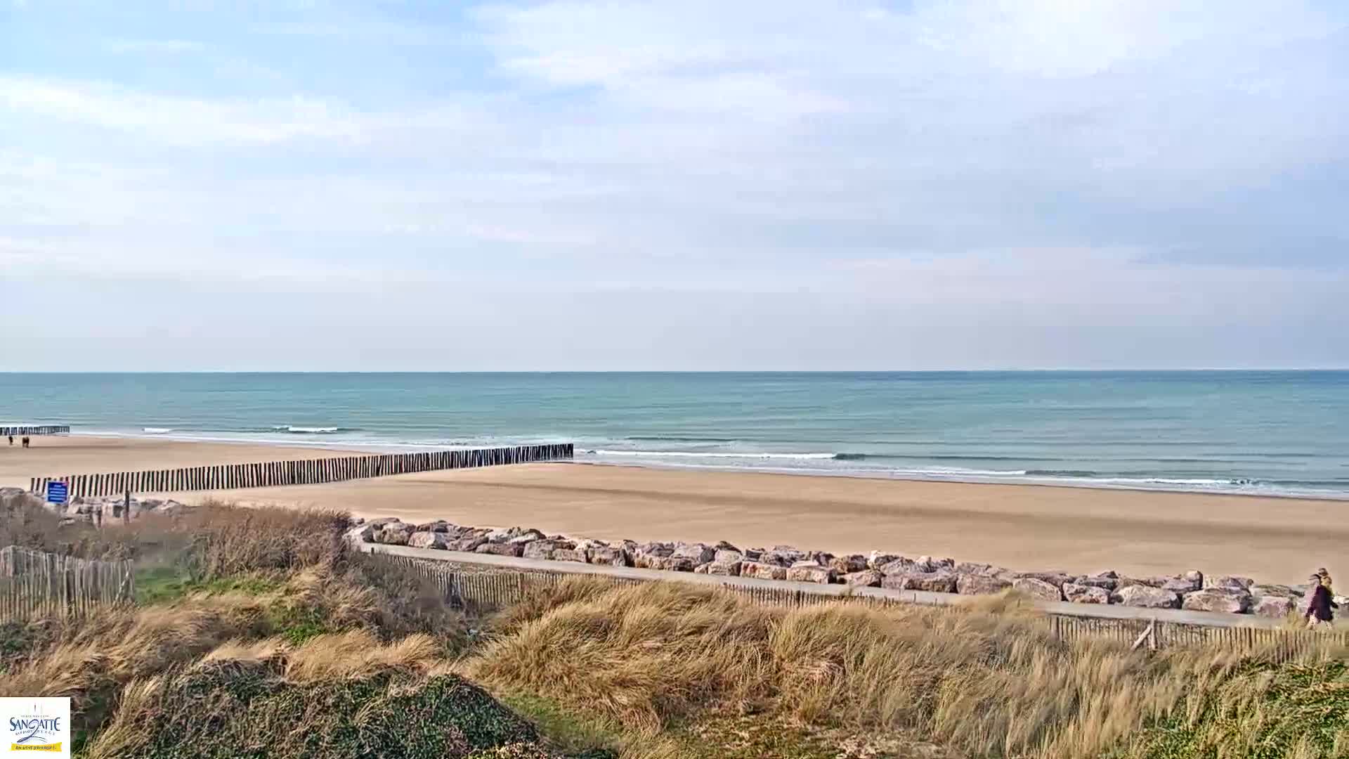 A sandy beach with calm ocean waves under a partly cloudy sky is bordered by a rock wall and grassy dunes.