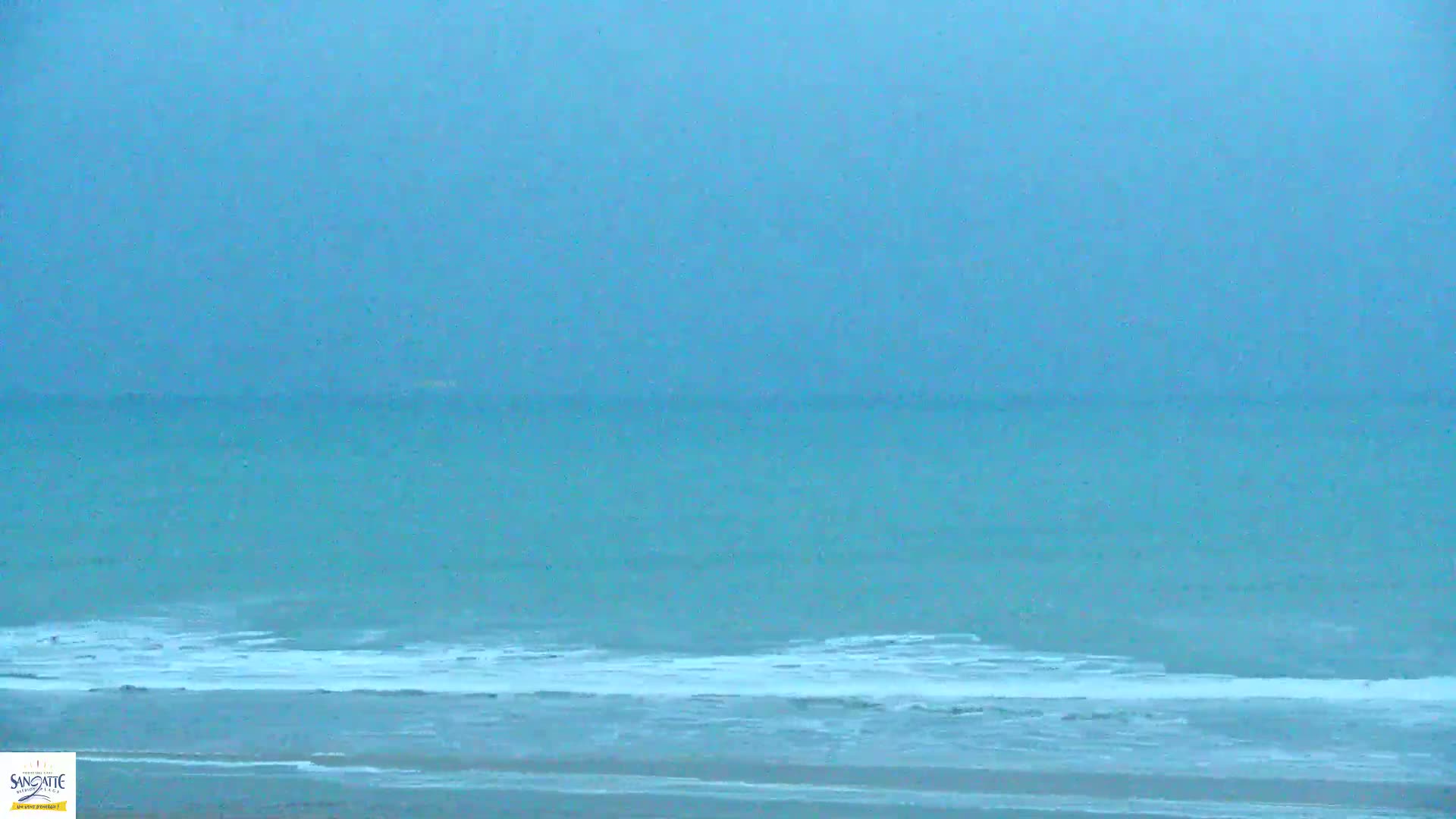 A hazy, overcast sky hangs over a calm, teal ocean with small waves breaking on a sandy beach.
