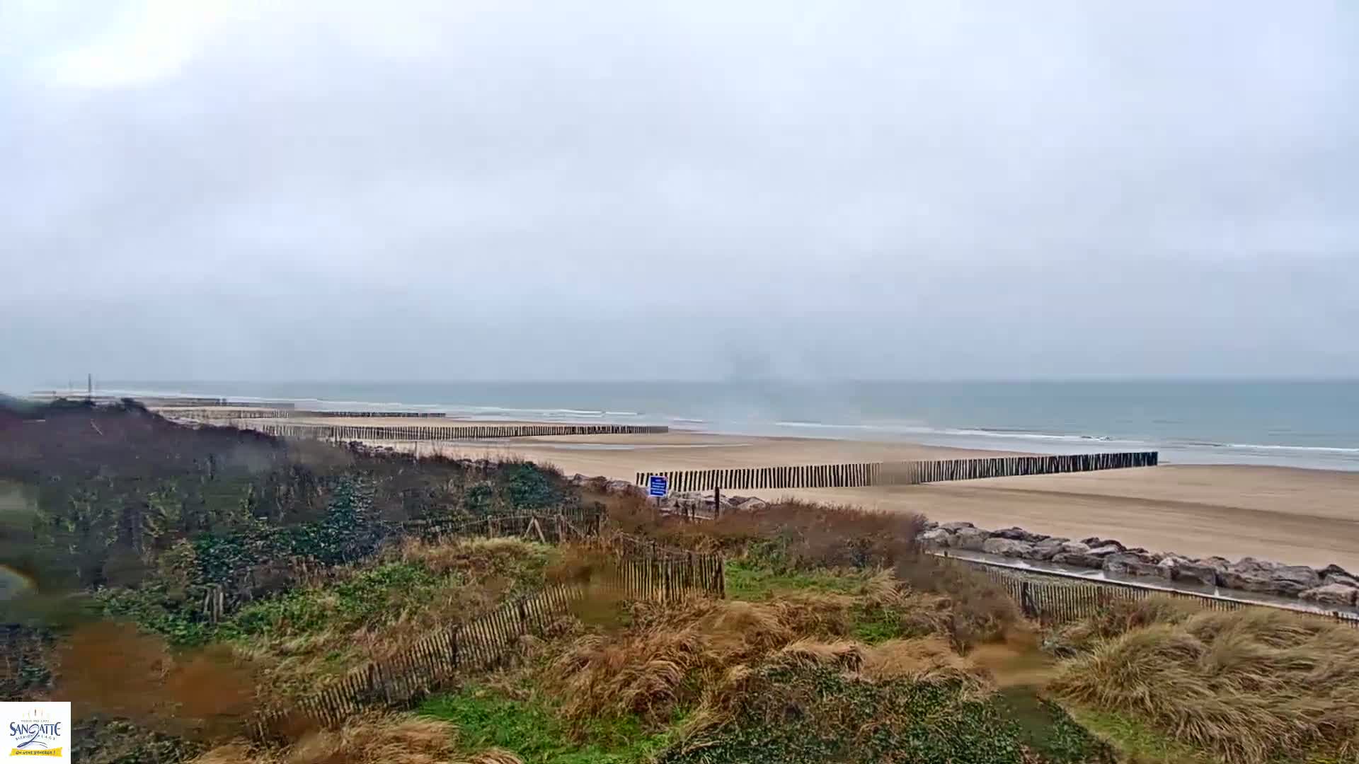 A sandy beach with low wooden barriers stretches along a calm, gray ocean under an overcast sky.
