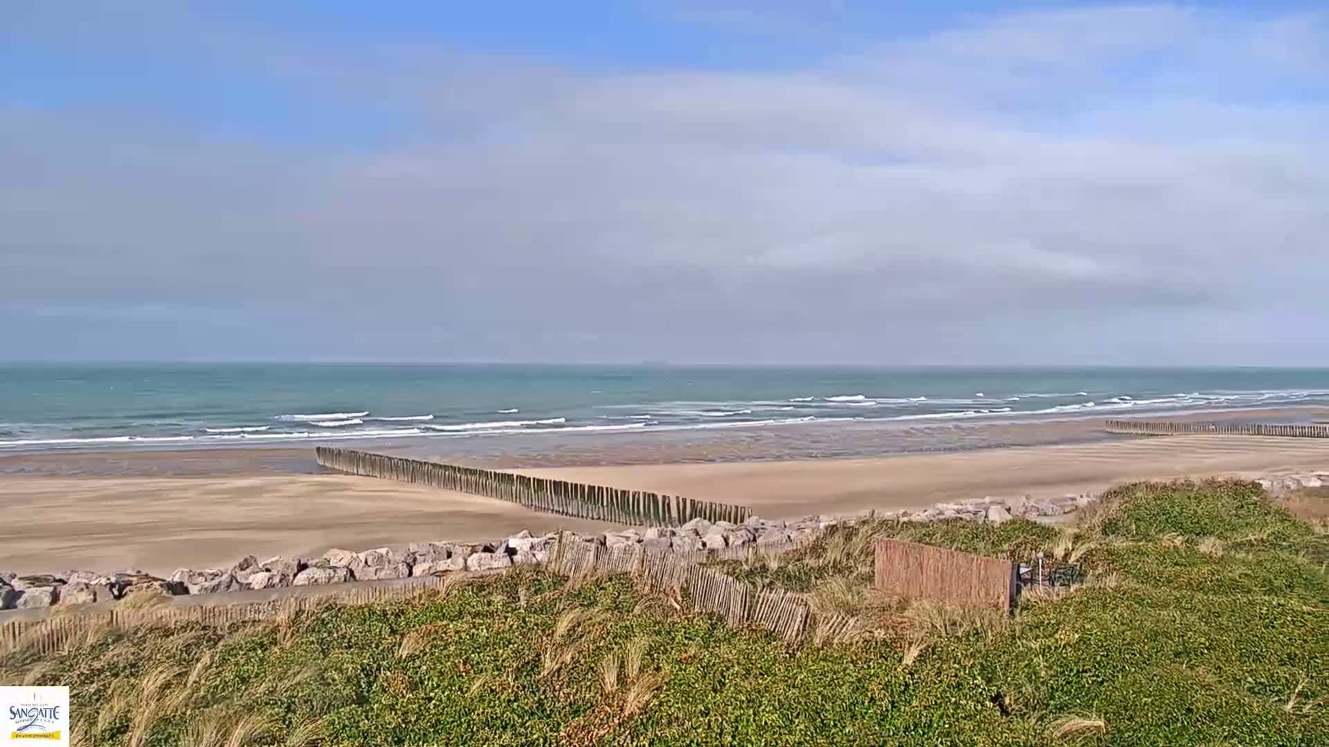 A sandy beach with gentle waves under a partly cloudy sky is protected by a long line of wooden posts.