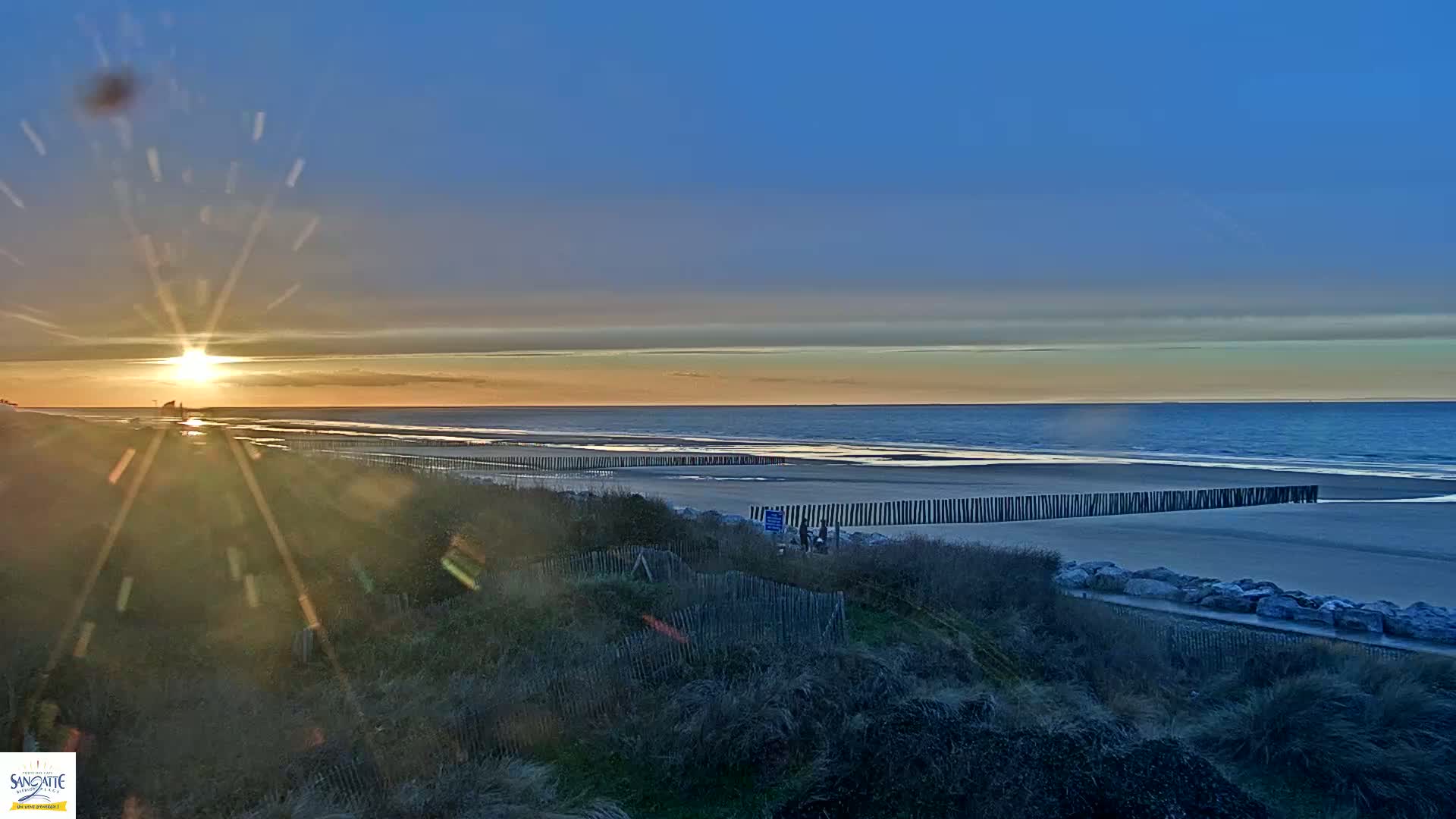 A sunset over a calm ocean beach, viewed from behind a dune with a wooden barrier along the shoreline.