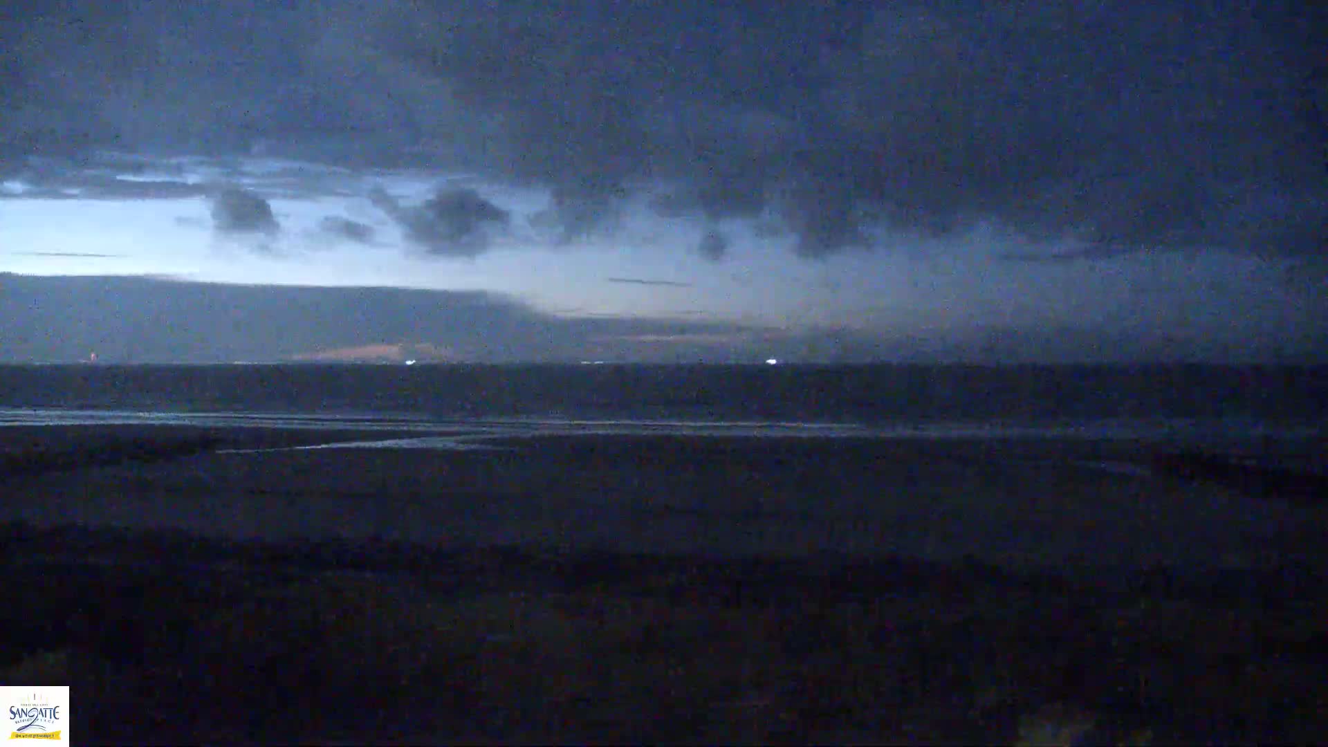 A dark, overcast sky hangs over a calm, dark ocean and a flat, dark beach at twilight.