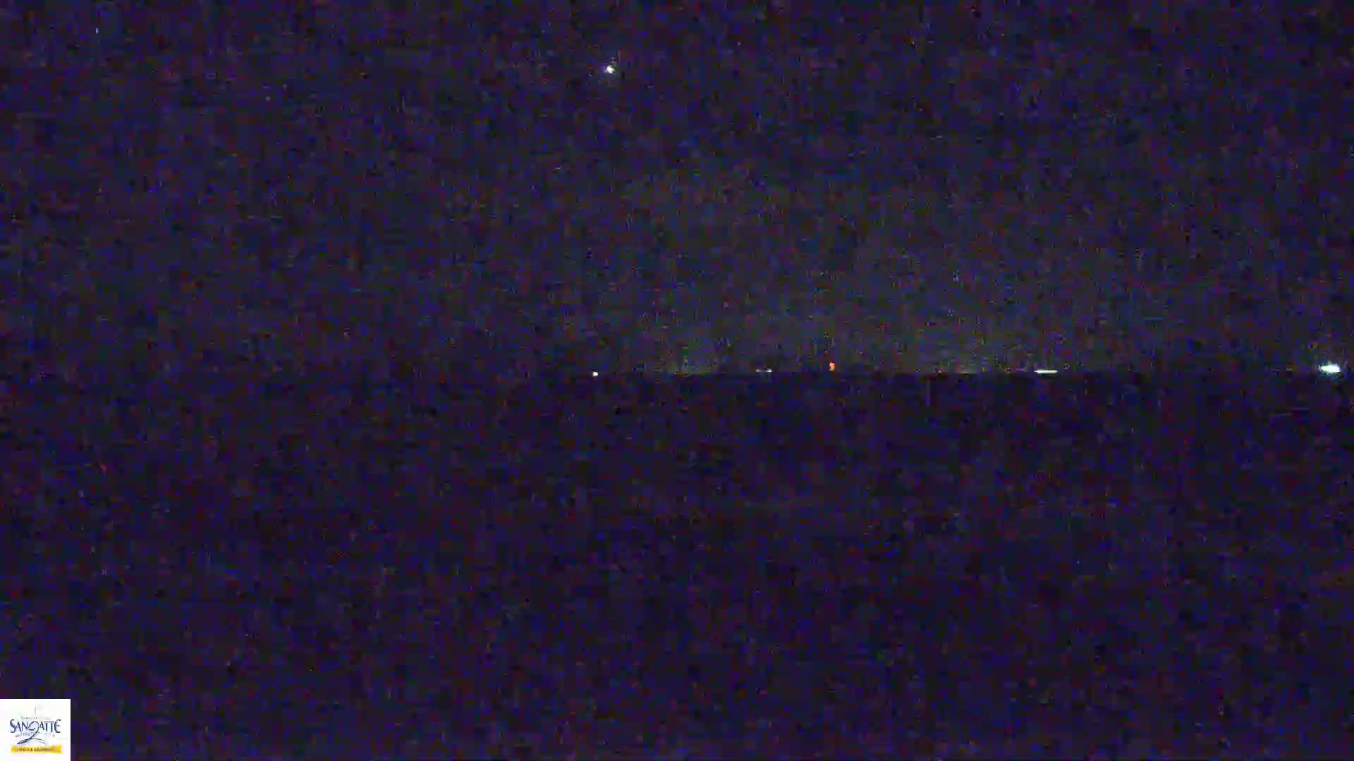 A dark, nighttime landscape shows a flat horizon with a few distant lights under a mostly clear, starry sky.