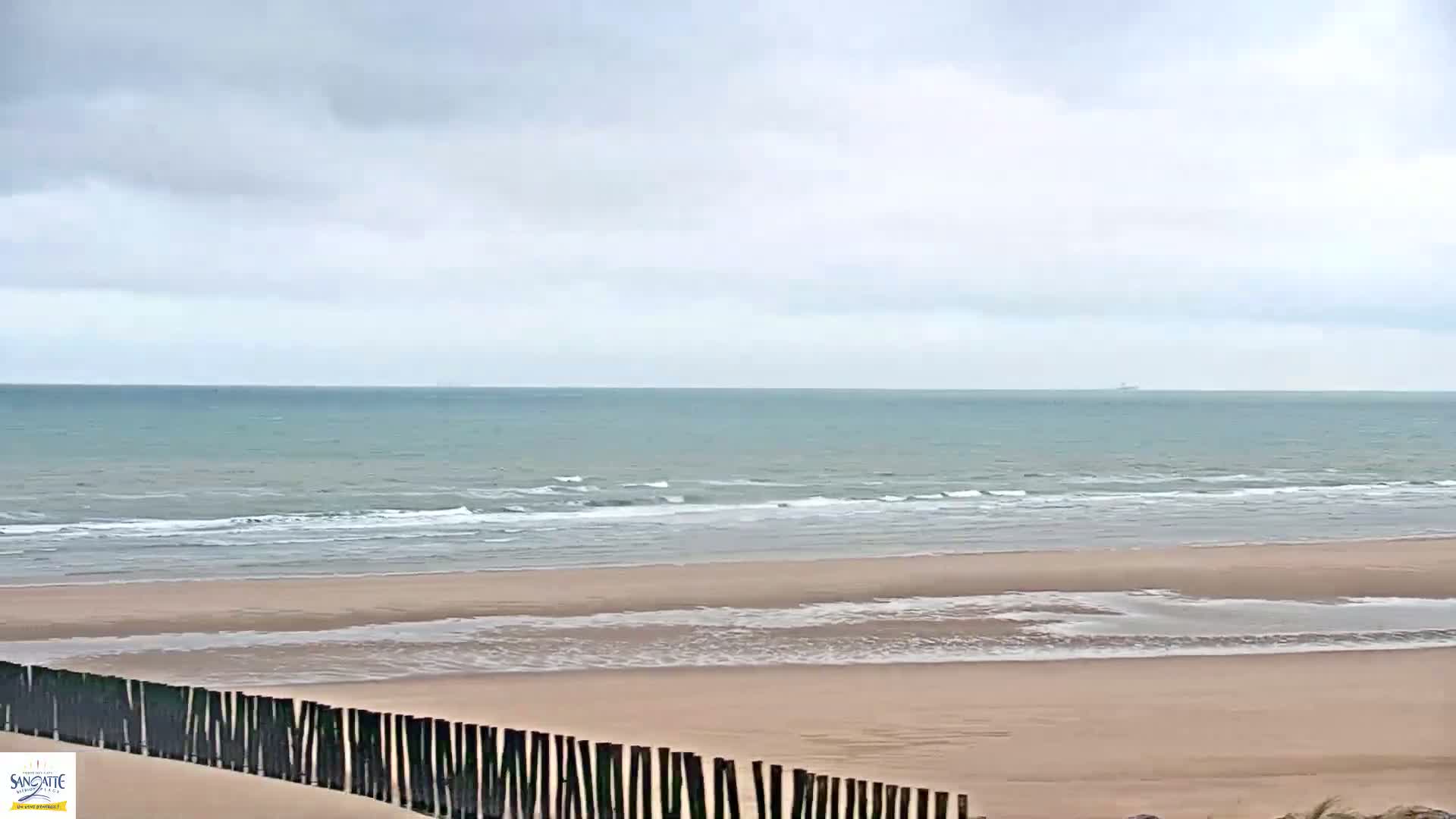 A sandy beach with small waves under a cloudy sky is partially obscured by a dark wooden fence.