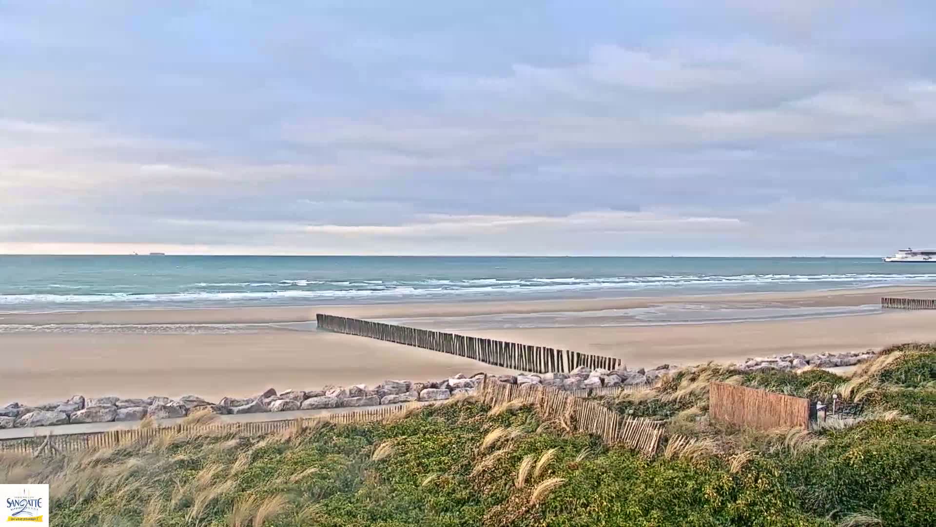 A sandy beach with a calm ocean under a cloudy sky is partially protected by a wooden fence and large rocks.