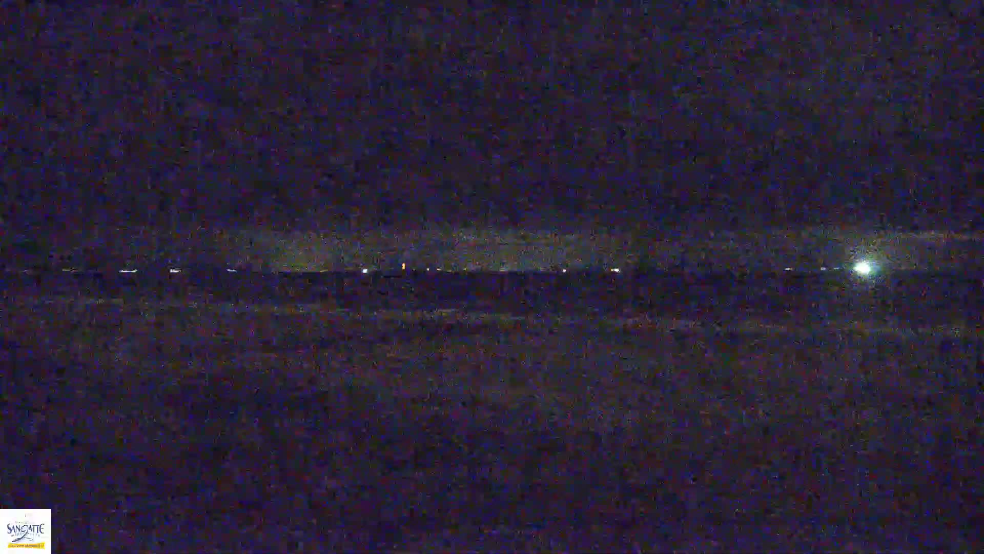 A nighttime, low-light view shows a dark, flat expanse of land with a distant line of faint lights under a dark sky.