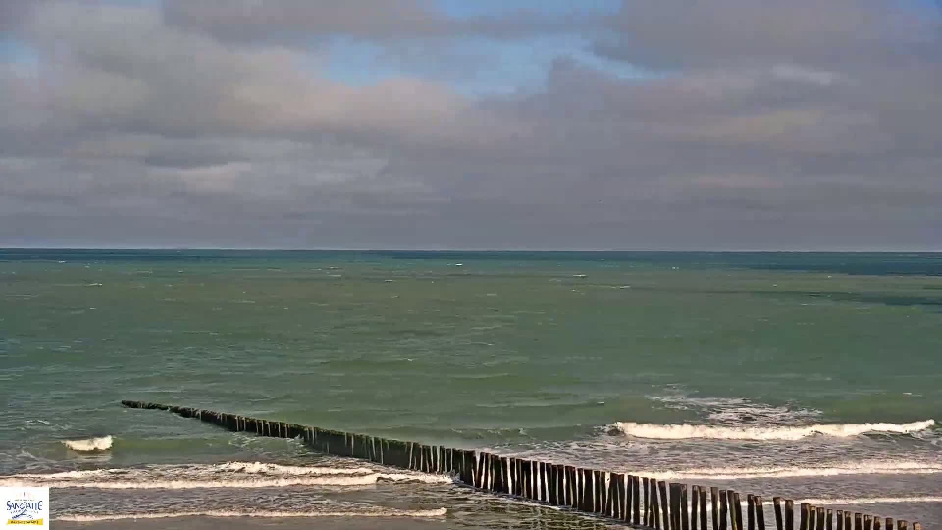 A partially cloudy sky overlooks a greenish-blue sea with small waves breaking on a beach protected by a long, low wooden breakwater.