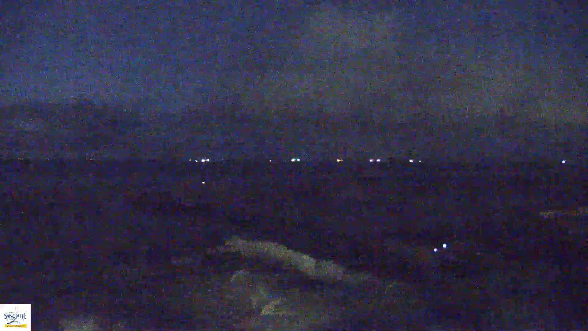 A dark, nighttime scene shows a distant line of faint lights across a dark body of water under a mostly clear, dark blue sky.