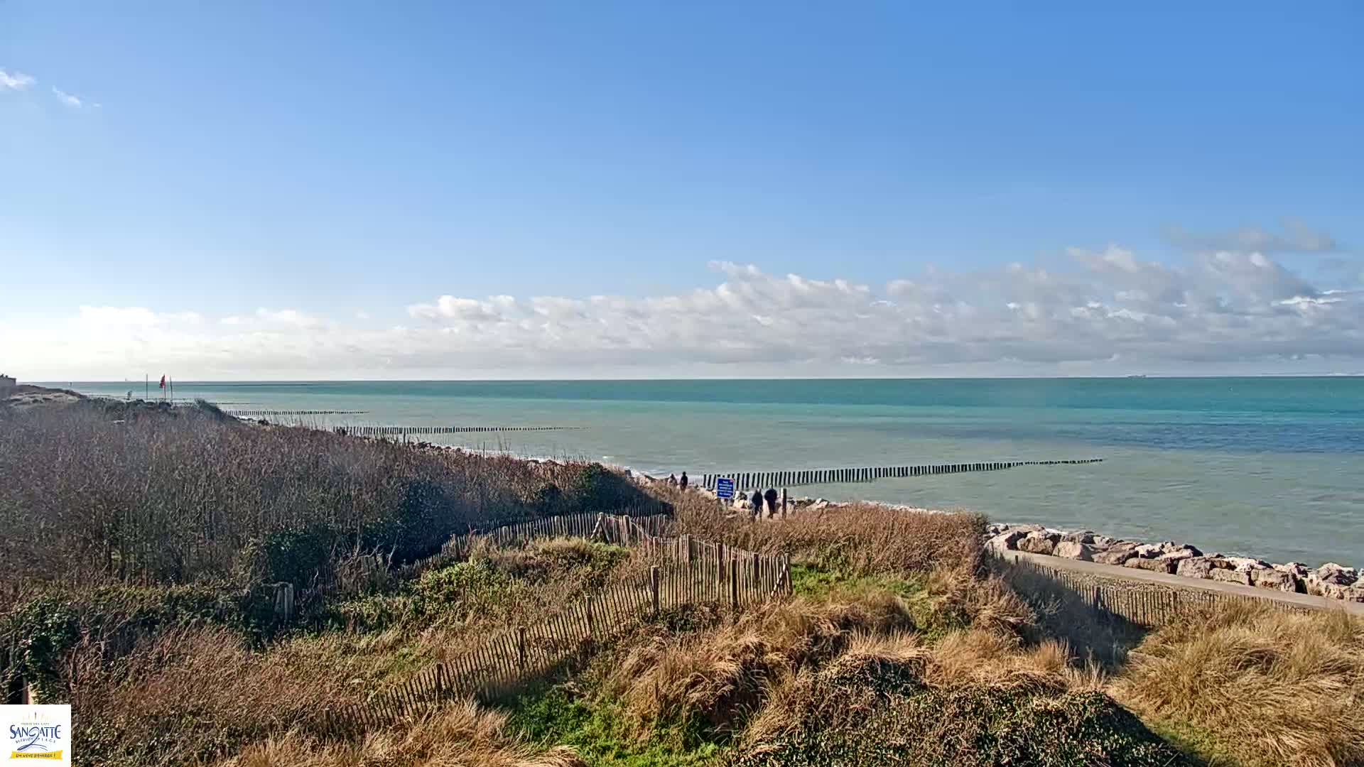 A partly cloudy day reveals a calm teal sea with a rocky shore and dune vegetation, where several people walk along a path.