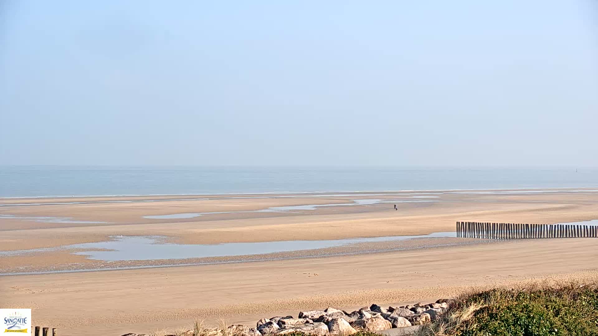 A wide expanse of sandy beach with shallow pools of water stretches to a calm ocean under a clear, light blue sky.
