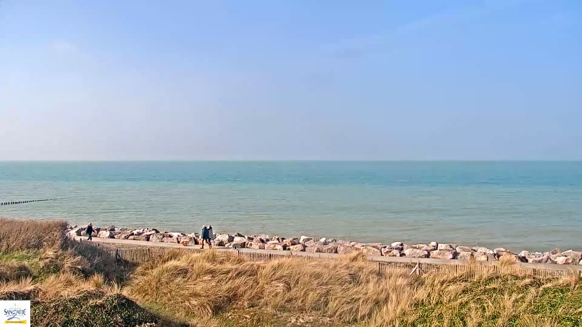 A calm, blue-green sea stretches to the horizon under a clear blue sky, with a few people walking along a path lined with rocks and bordered by sandy dunes.