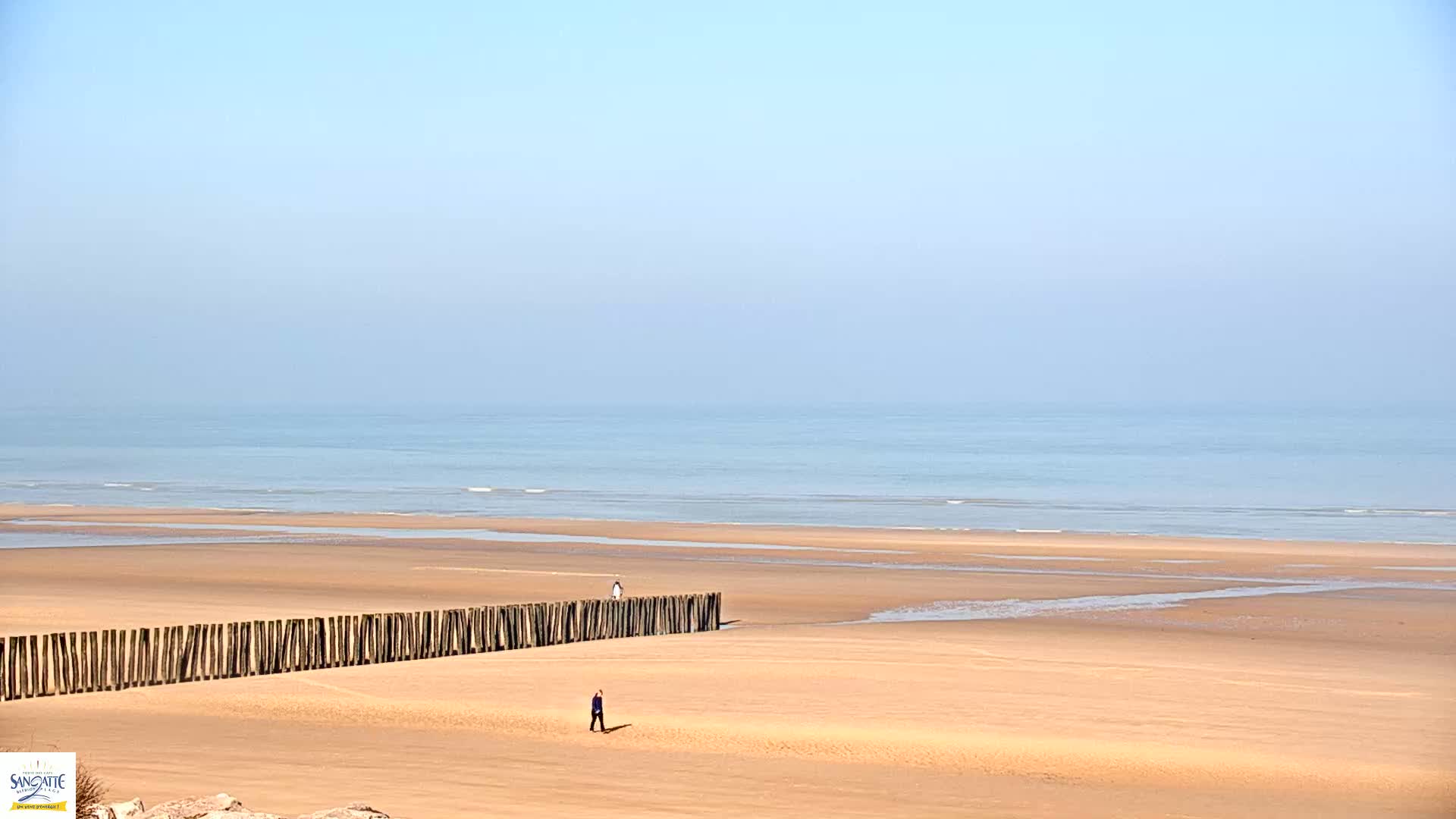 A long, sandy beach stretches to a calm, light blue ocean under a clear, pale blue sky; a wooden barrier runs parallel to the water's edge, and two lone figures walk on the sand.
