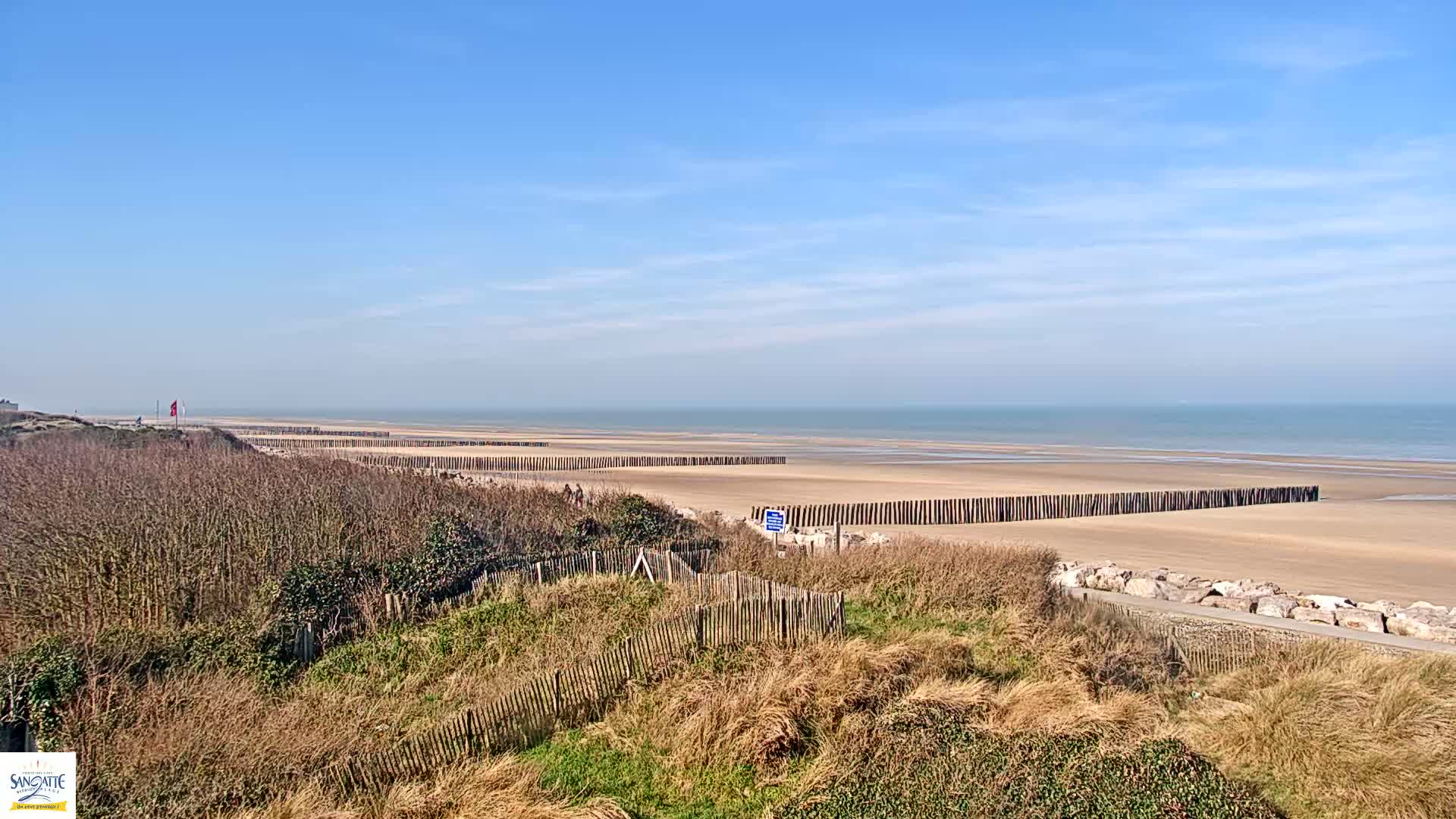 A sandy beach with wooden posts along the shoreline is visible from a dune covered in dried grasses under a clear blue sky.
