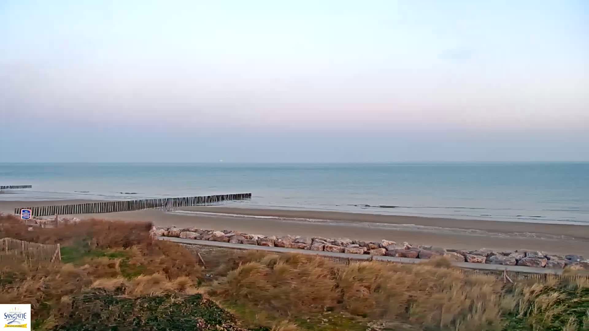 A calm, pale-blue sea stretches to the horizon under a pale, clear sky, meeting a wide sandy beach protected by rows of wooden posts and large rocks.